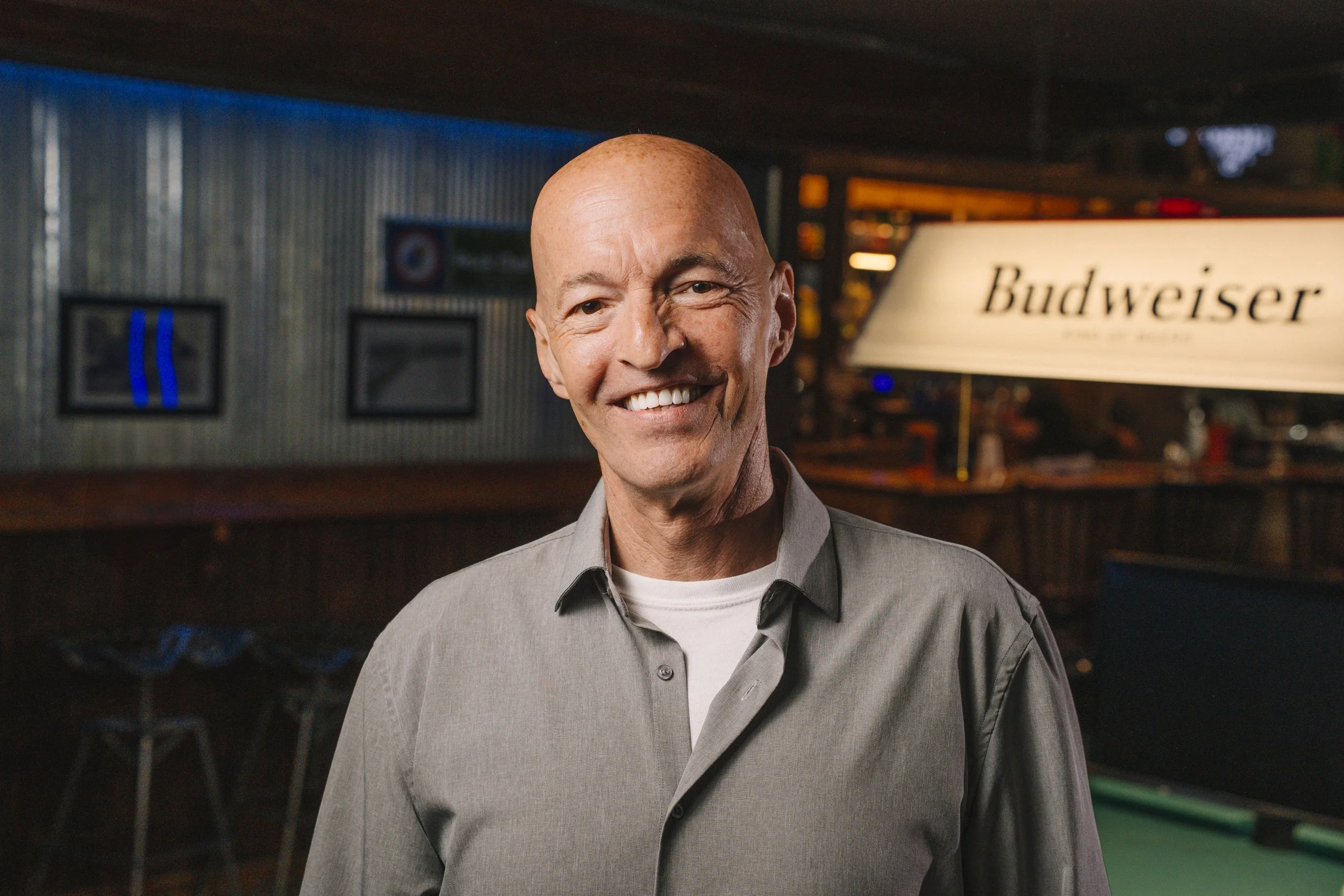 Smiling man with a bald head in a gray shirt in a bar or pub setting, with a Budweiser sign in the background.
