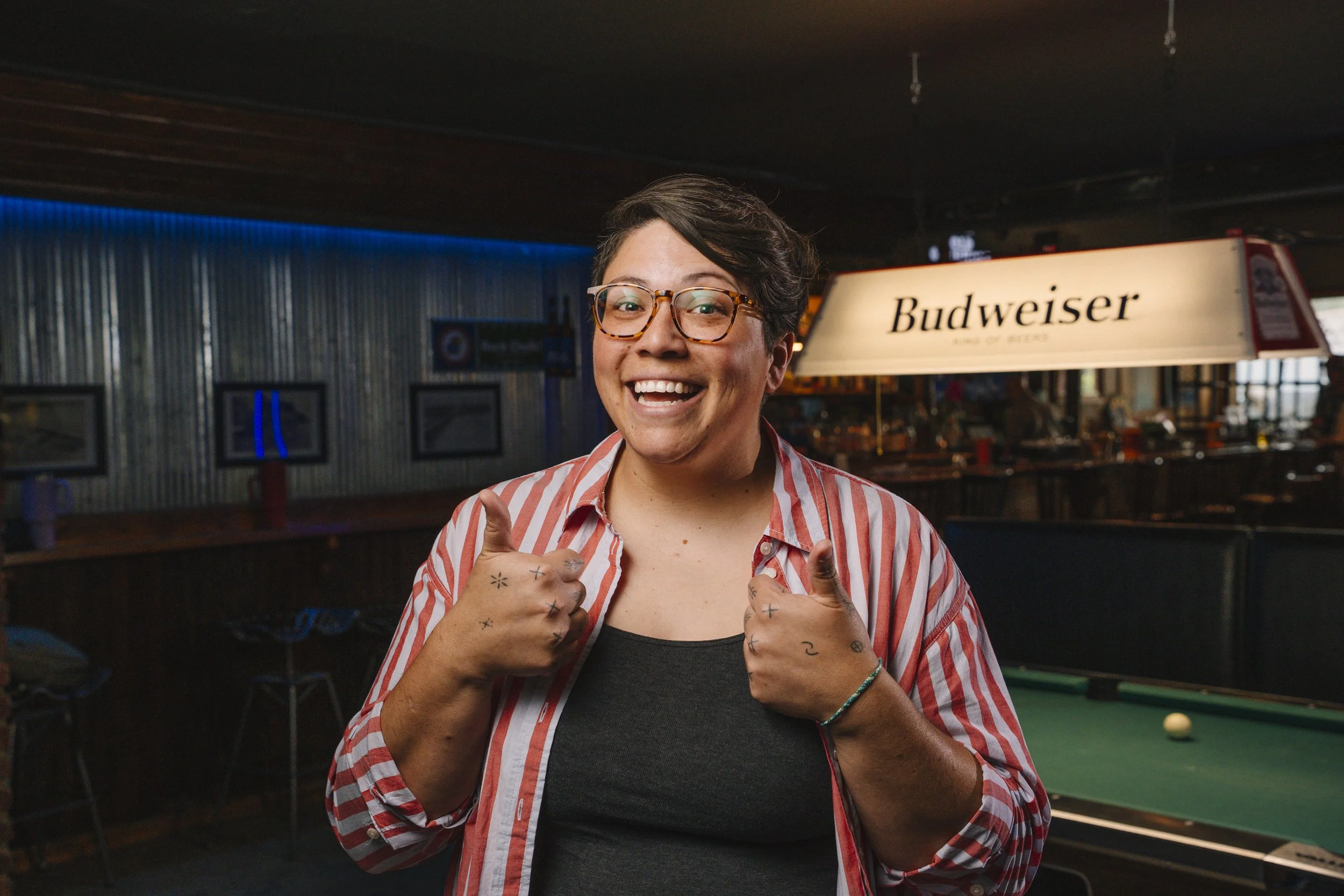 A person with short dark hair, glasses, and tattoos on their hands smiling and giving a thumbs up in a bar setting with a Budweiser sign and pool table in the background.