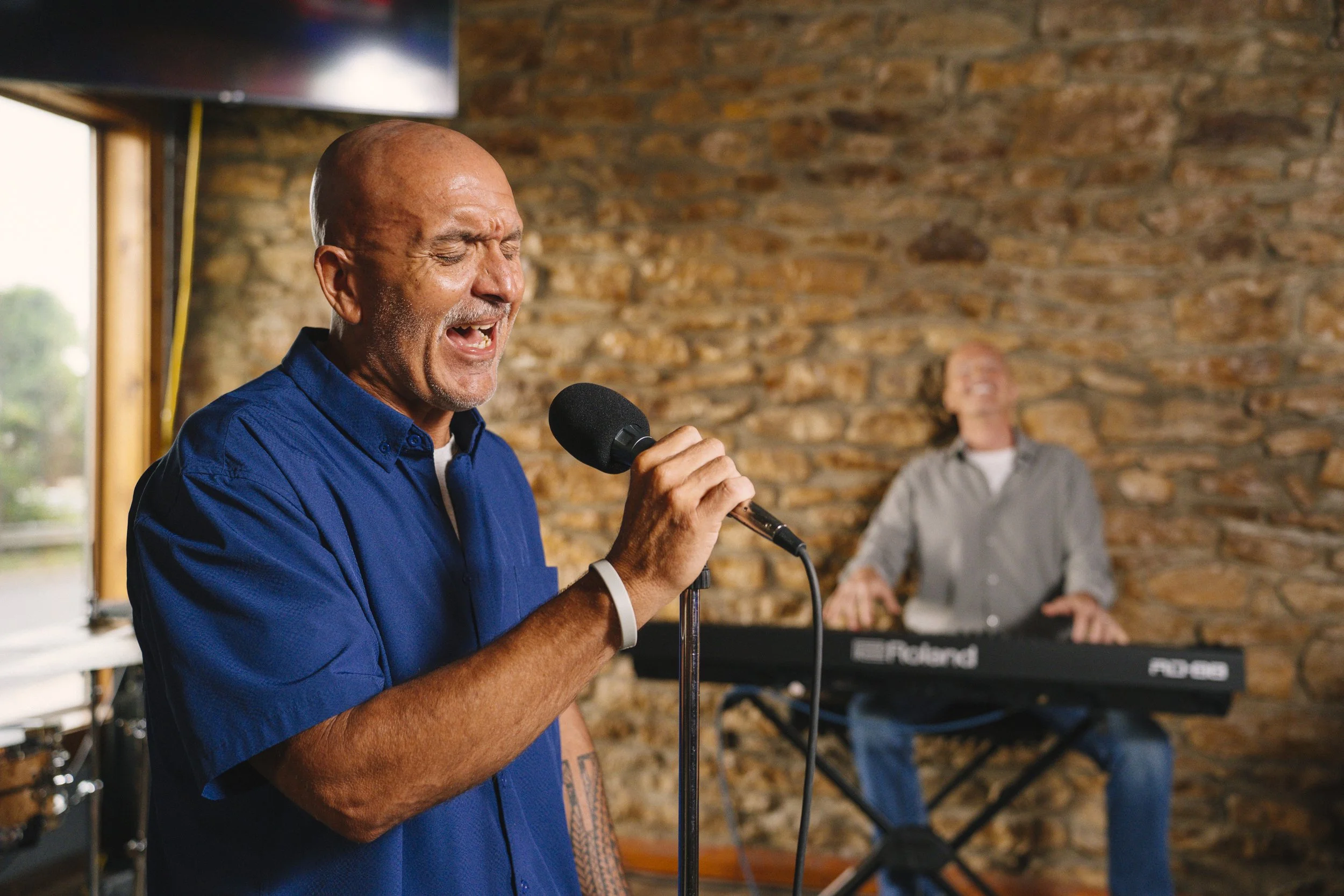 An elderly man passionately singing into a microphone during a live performance, with a keyboard player in the background against a brick wall.