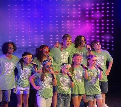 A group of children and a young adult singing on stage with microphones, all wearing matching green t-shirts with the text 'Our Voices Change The World,' under purple and pink stage lighting.