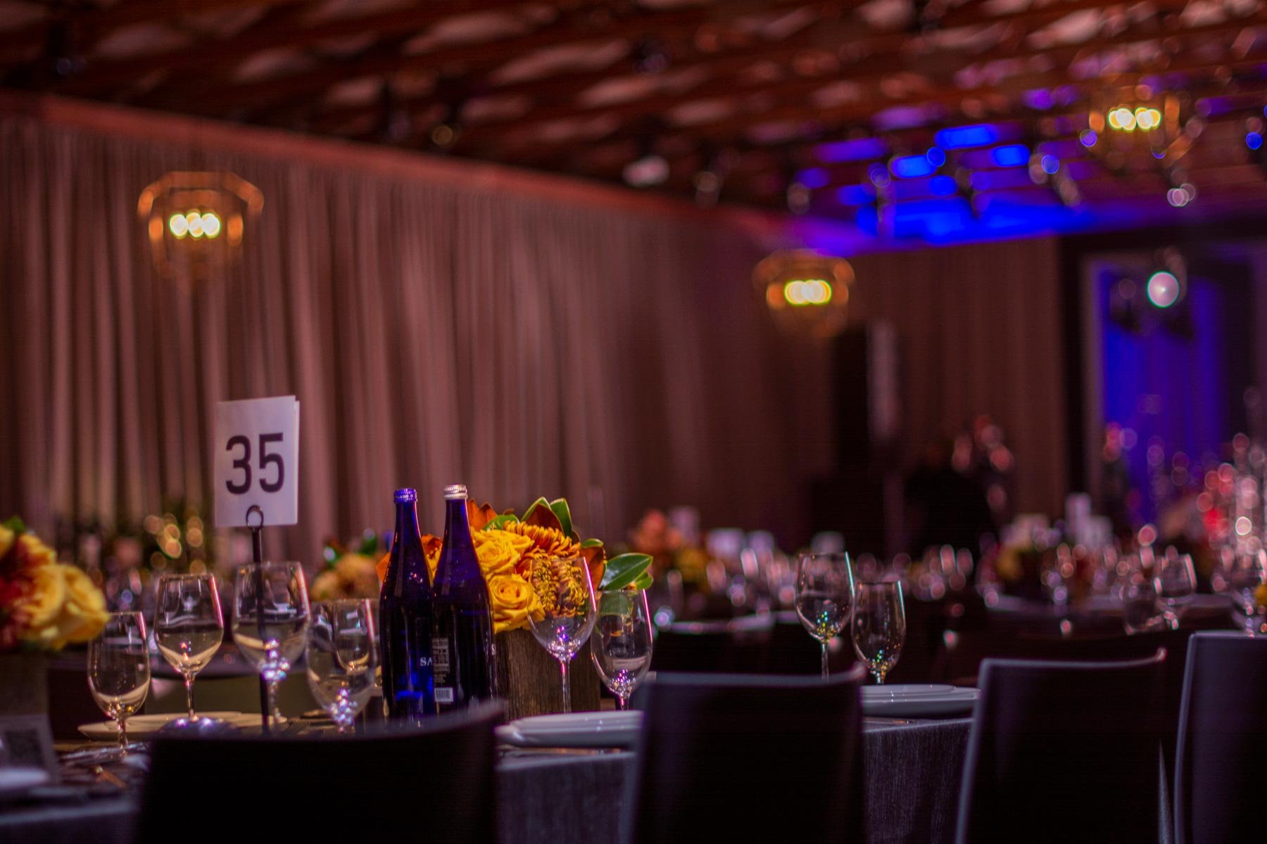 Elegant banquet table setting with flower centerpieces, wine glasses, water bottles, and a table number card, in a dimly lit event hall with colorful lighting.