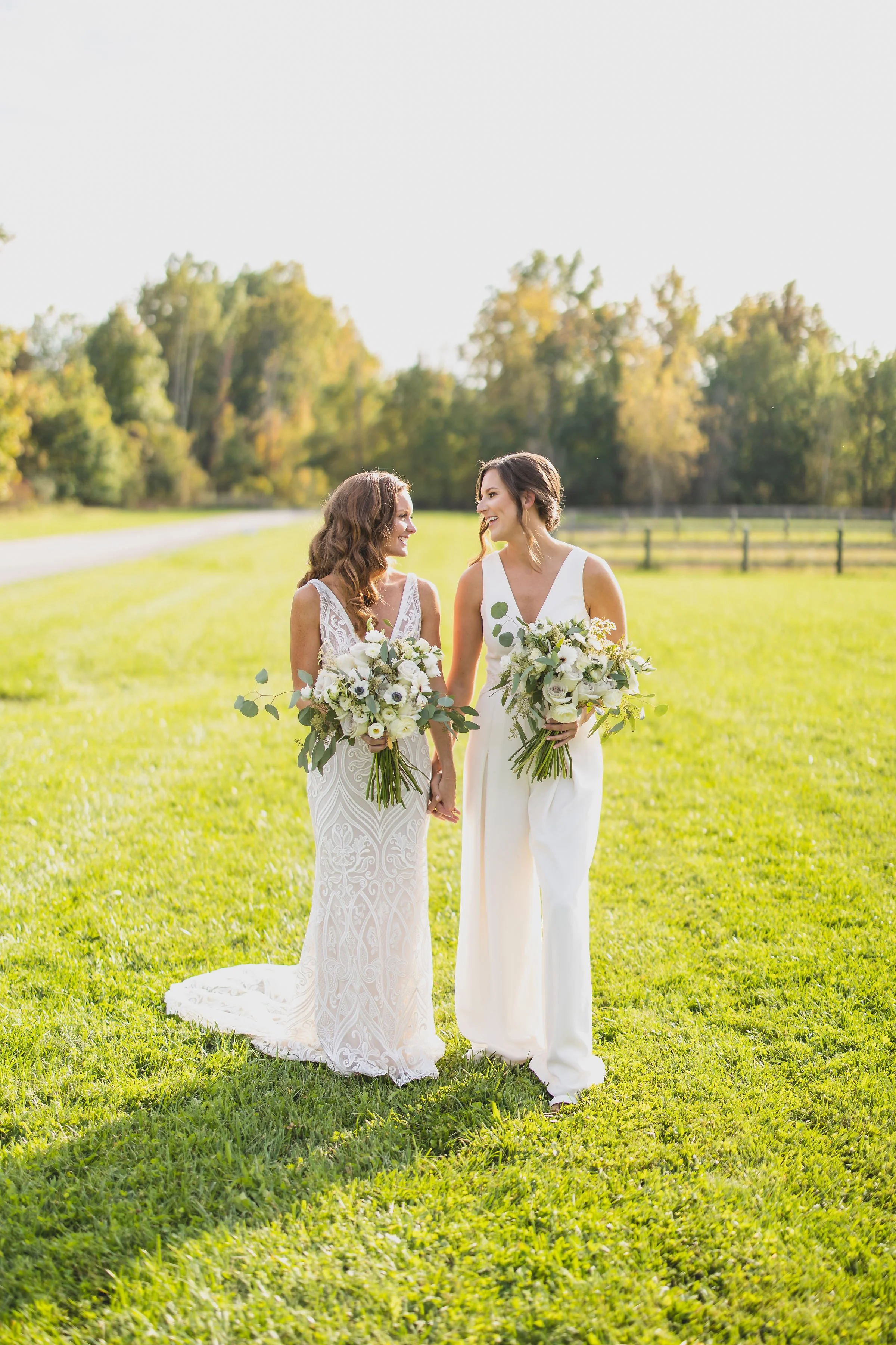 Two women in wedding dresses holding bouquets, smiling and holding hands outdoors on a sunny day with green grass and trees in the background.
