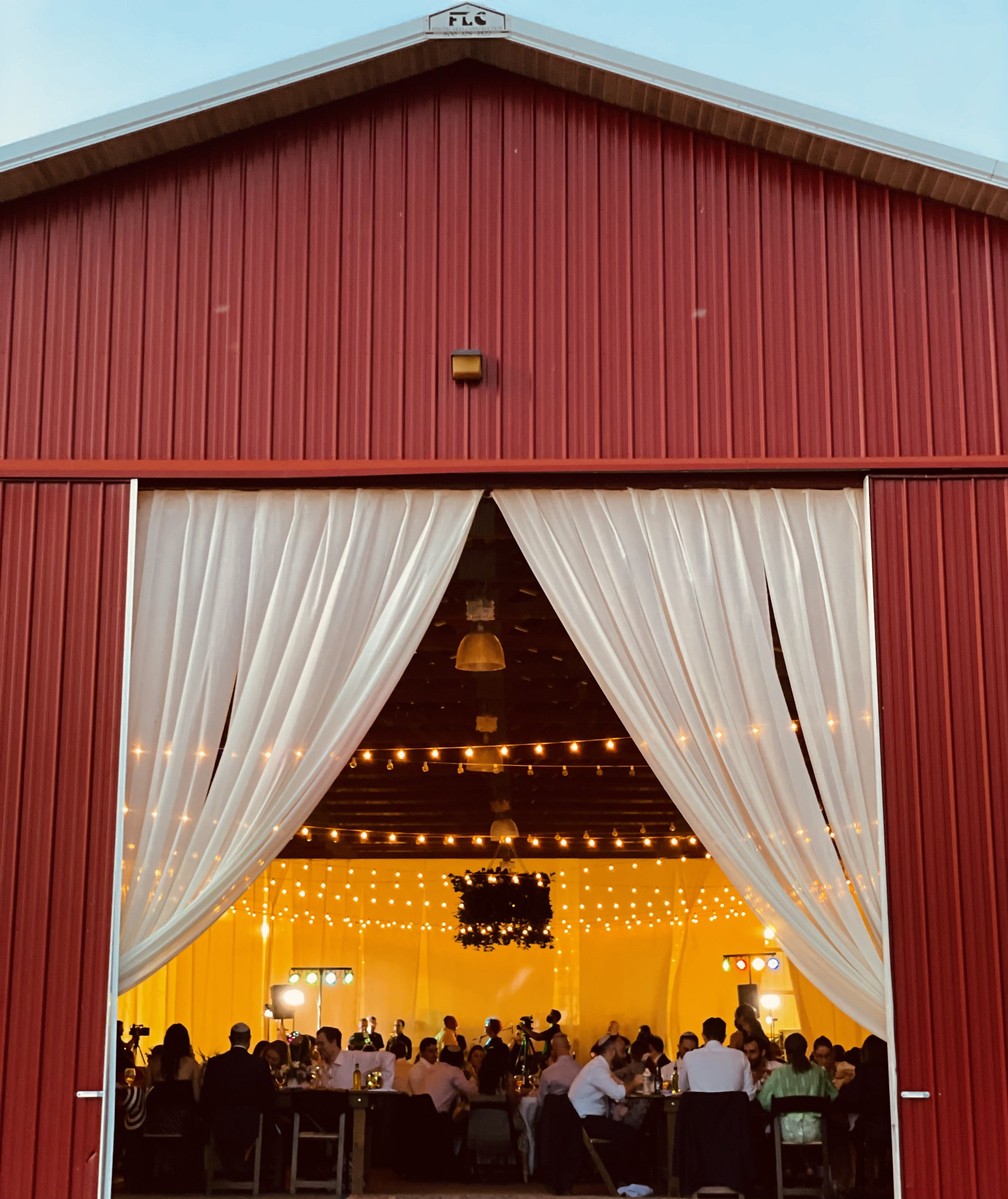 A view of a lively indoor event space viewed through open barn doors, decorated with string lights and filled with people seated at tables, with a large hanging decoration in the center.