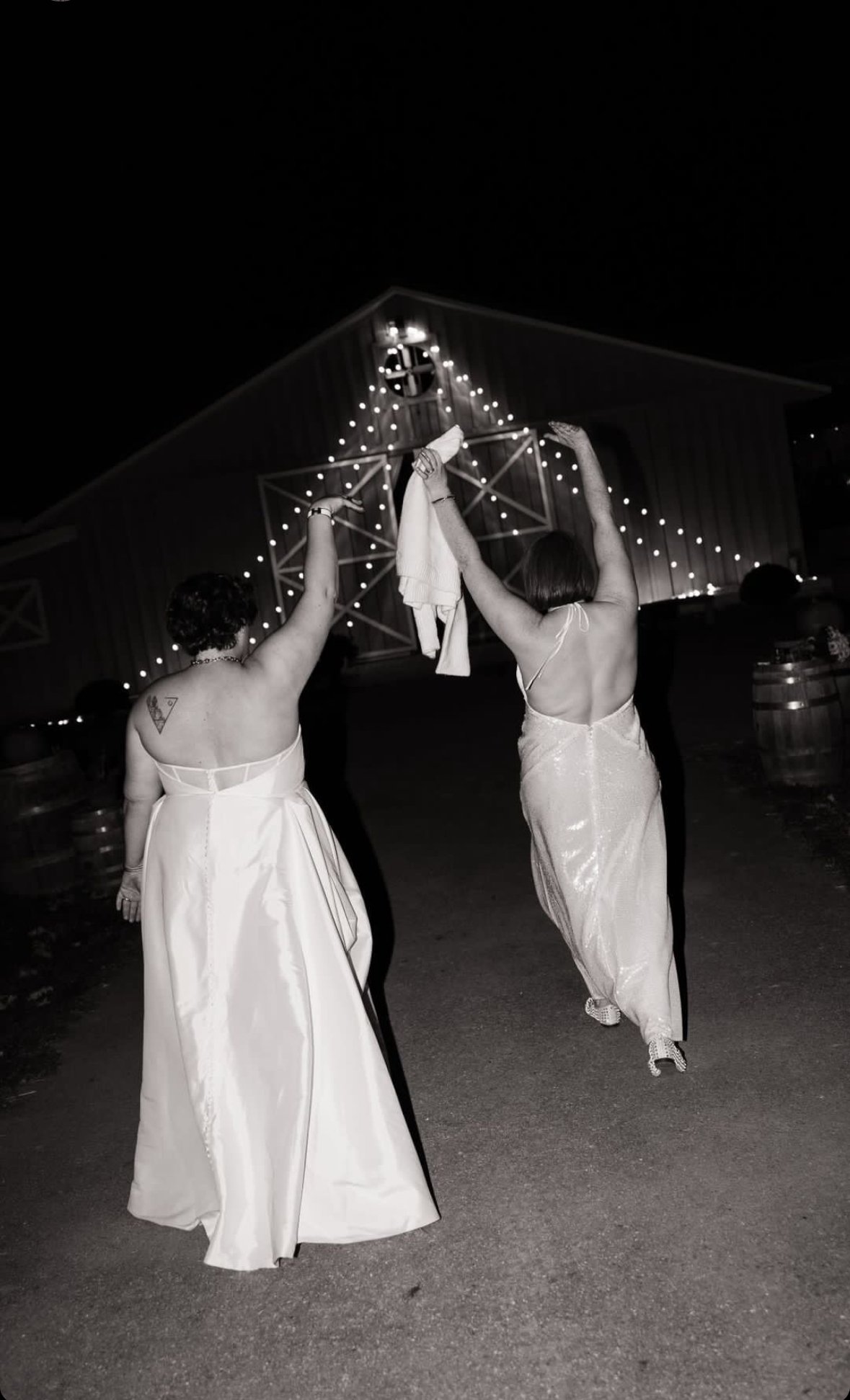 Two women in dresses walking away from the camera at night, holding clothes or a towel up in the air with a barn decorated with string lights in the background.