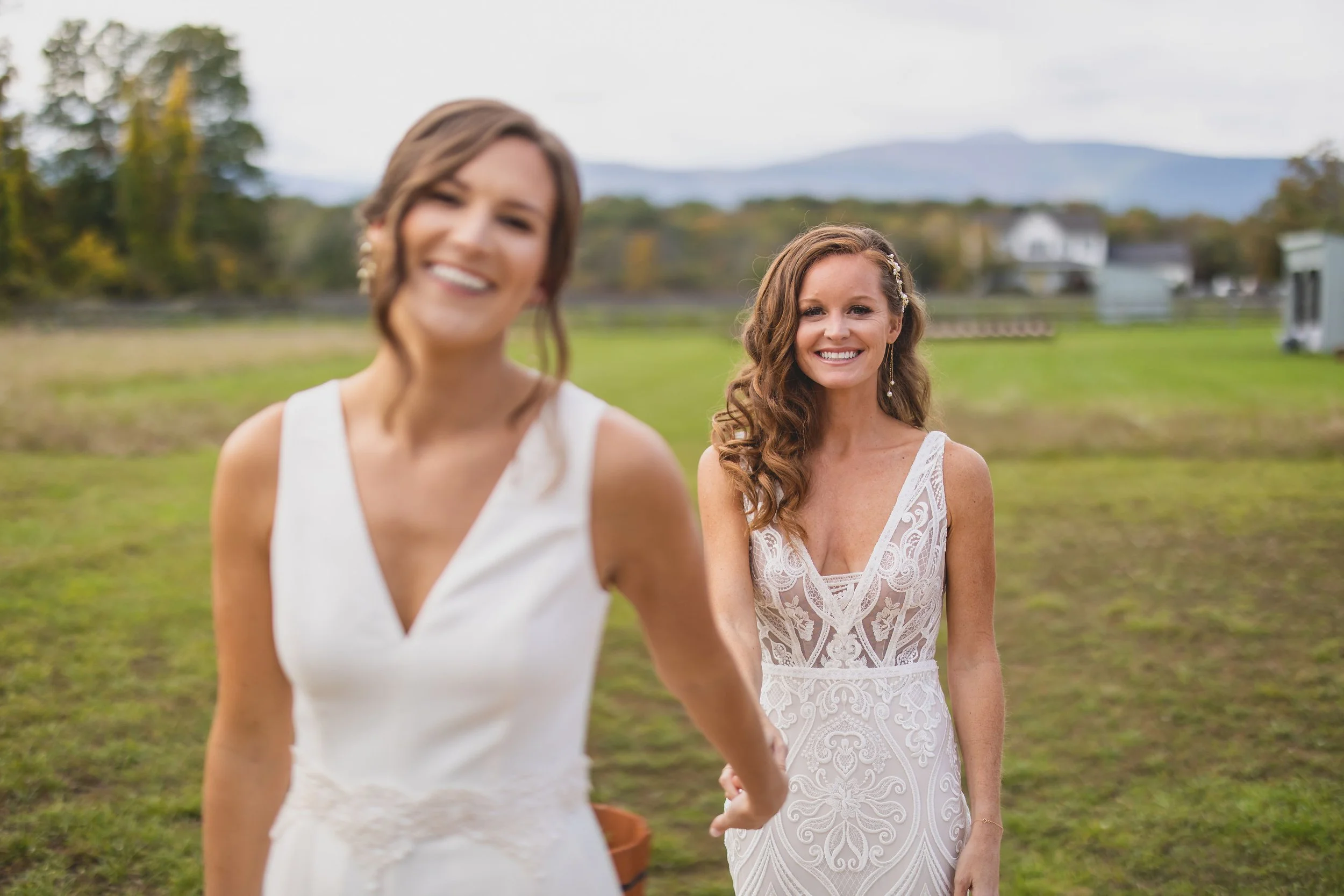 Two women in wedding dresses smiling outdoors on a grassy field with trees and mountains in the background.