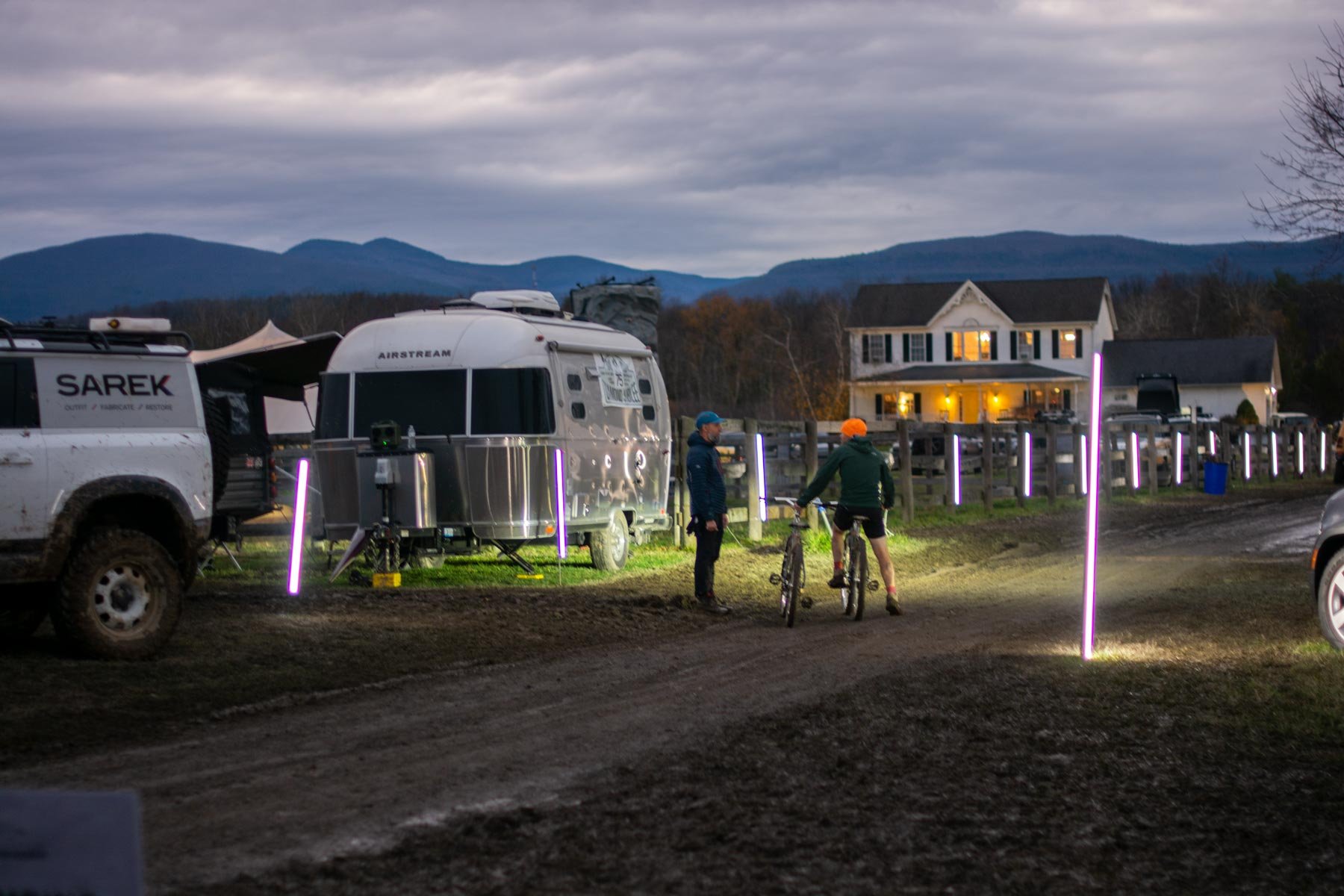 Two people stand near bicycles and a camper trailer on a dirt road, with a house in the background and mountains in the distance. The scene appears to be in the evening or dawn, with gathering lights and cloudy sky.