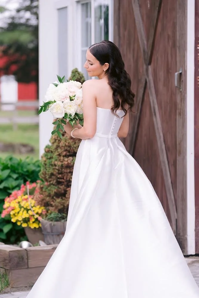 A bride in a strapless white wedding gown holding a bouquet of white flowers, standing outdoors near a wooden barn door, with colorful flowers and greenery around.