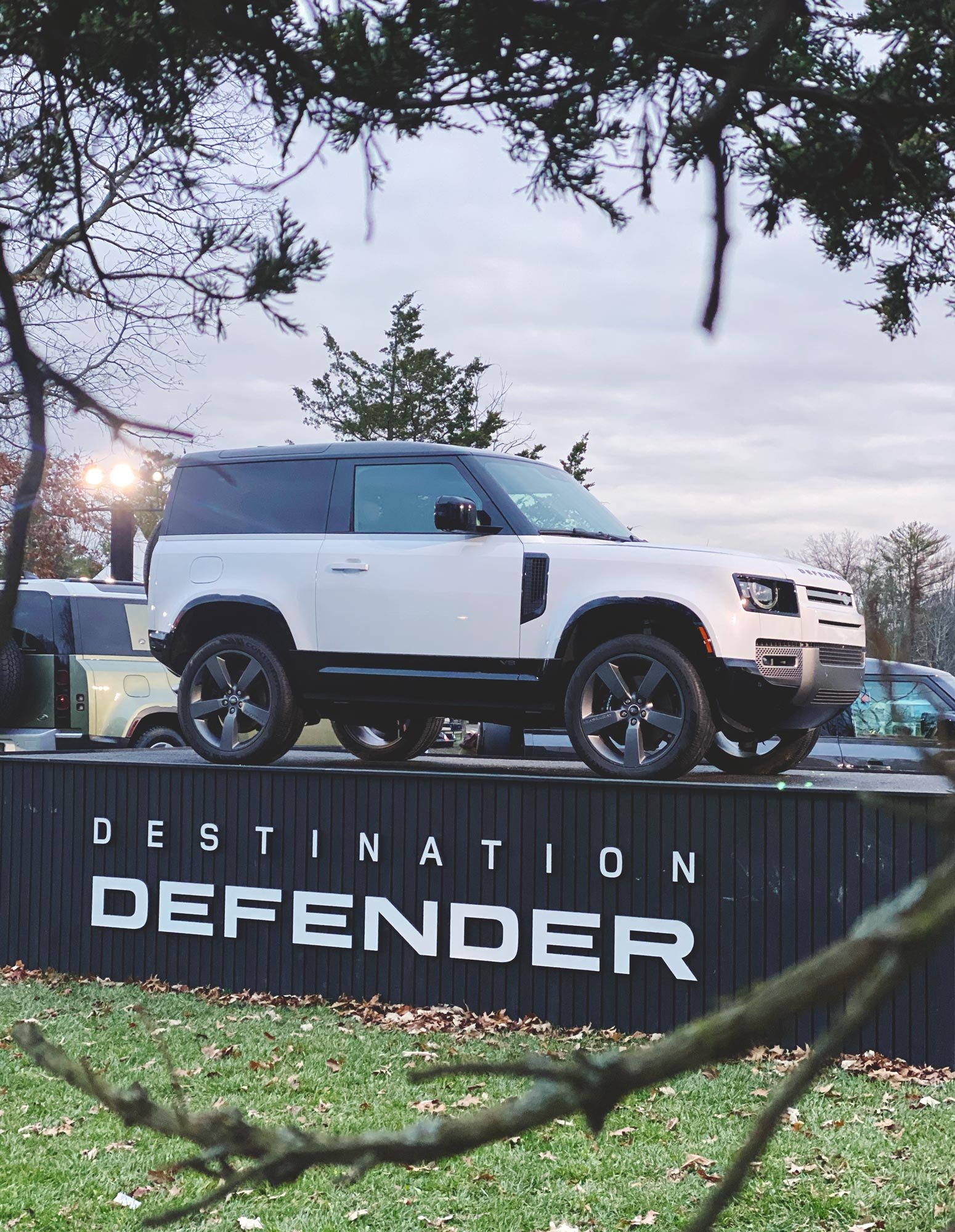 A white Range Rover SUV displayed on a black platform with the words 'Destination Defender' in white lettering. The background has trees and a cloudy sky, with some branches in the foreground.
