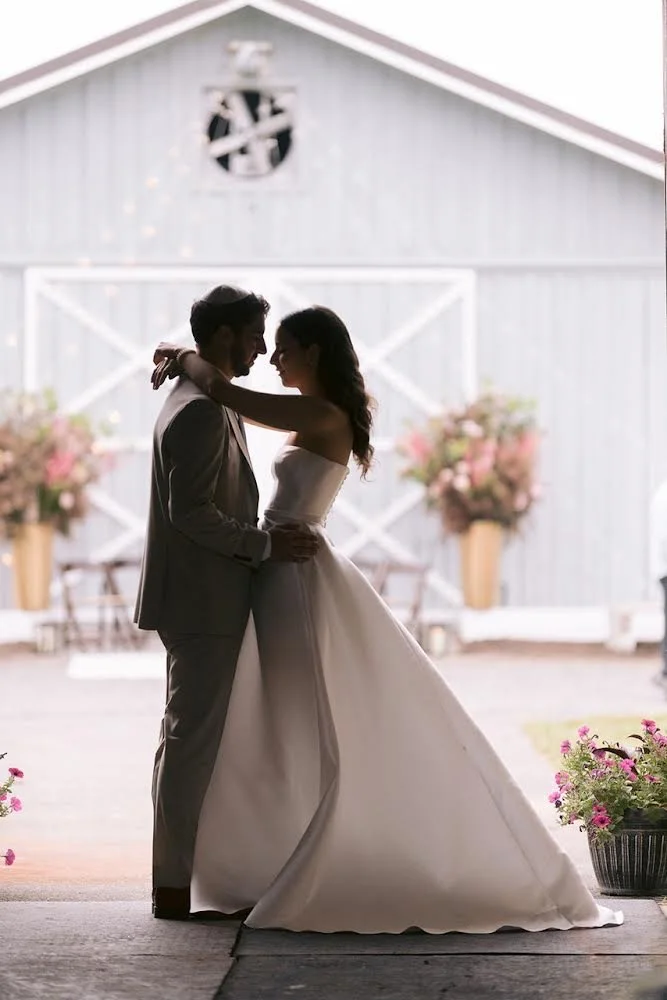 A bride and groom dancing in front of a barn decorated with pink flowers at a wedding reception.