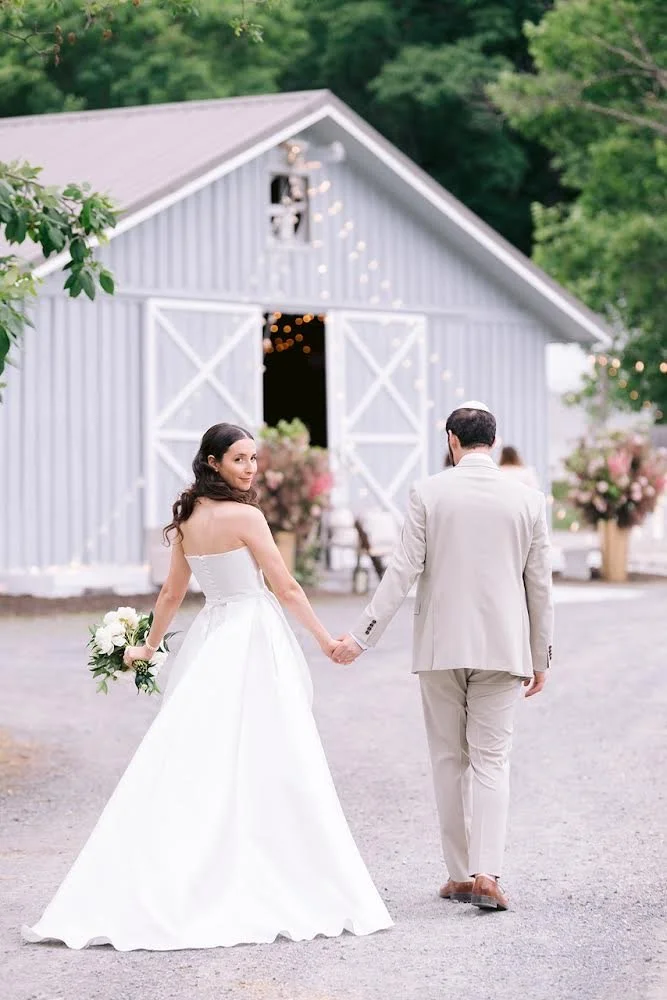 Bride and groom holding hands walking away from a barn decorated with string lights and flowers, on a wedding day.