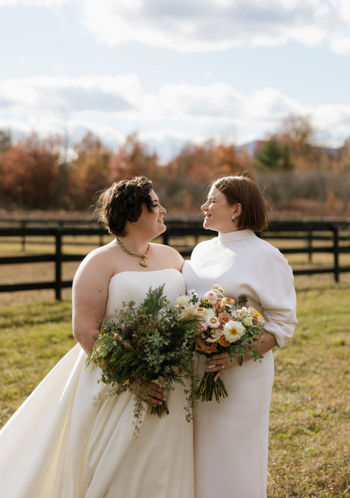 Two women dressed in wedding attire standing outdoors, holding bouquets of flowers, and smiling at each other with autumn trees and a black fence in the background.