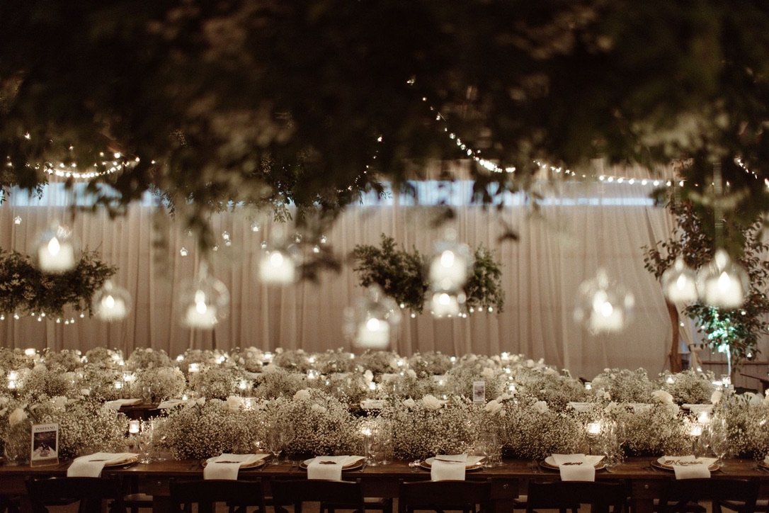 Decorated wedding reception table with white flowers, hanging lanterns, and string lights in a tented outdoor setting.