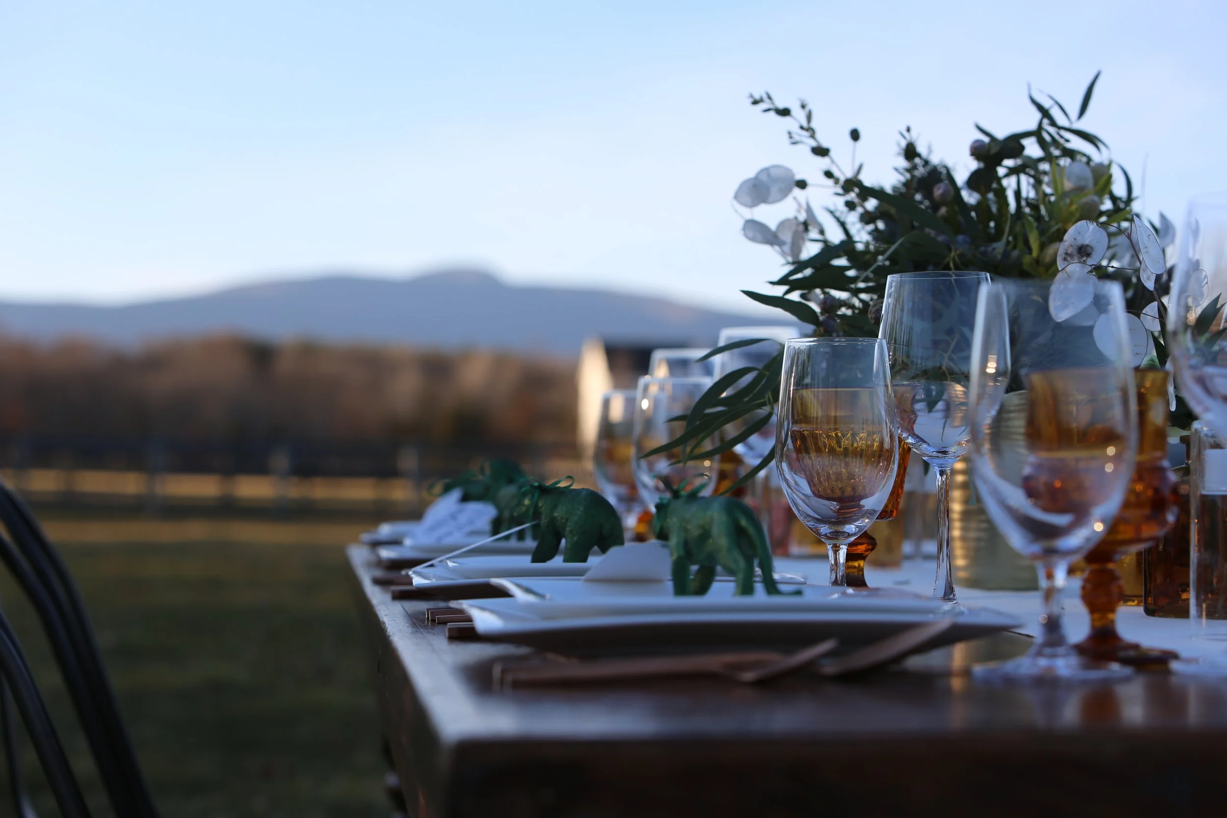 Outdoor dining table set with glasses, plates, and a floral centerpiece, with a mountain range in the background during evening.