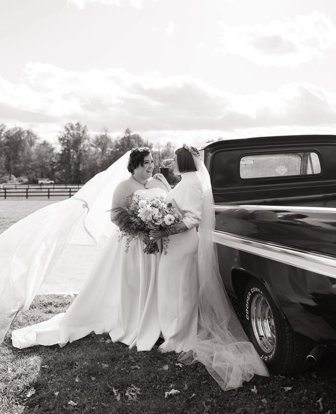 Two brides in wedding dresses share an emotional moment next to a vintage black pickup truck outdoors, with a field and trees in the background.