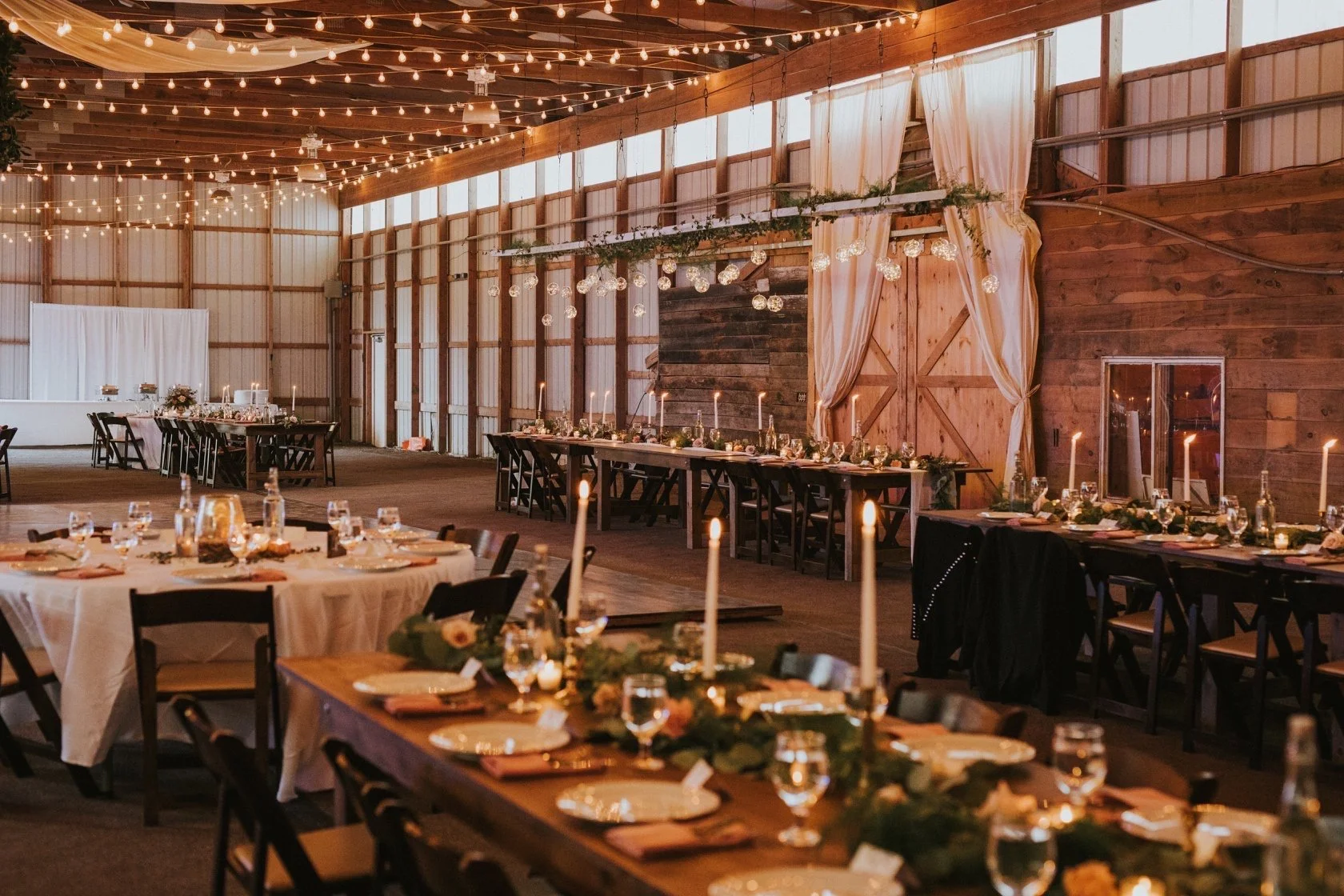 Interior of a rustic wooden hall decorated for a wedding reception with string lights, candlelit tables, and elegant table settings.