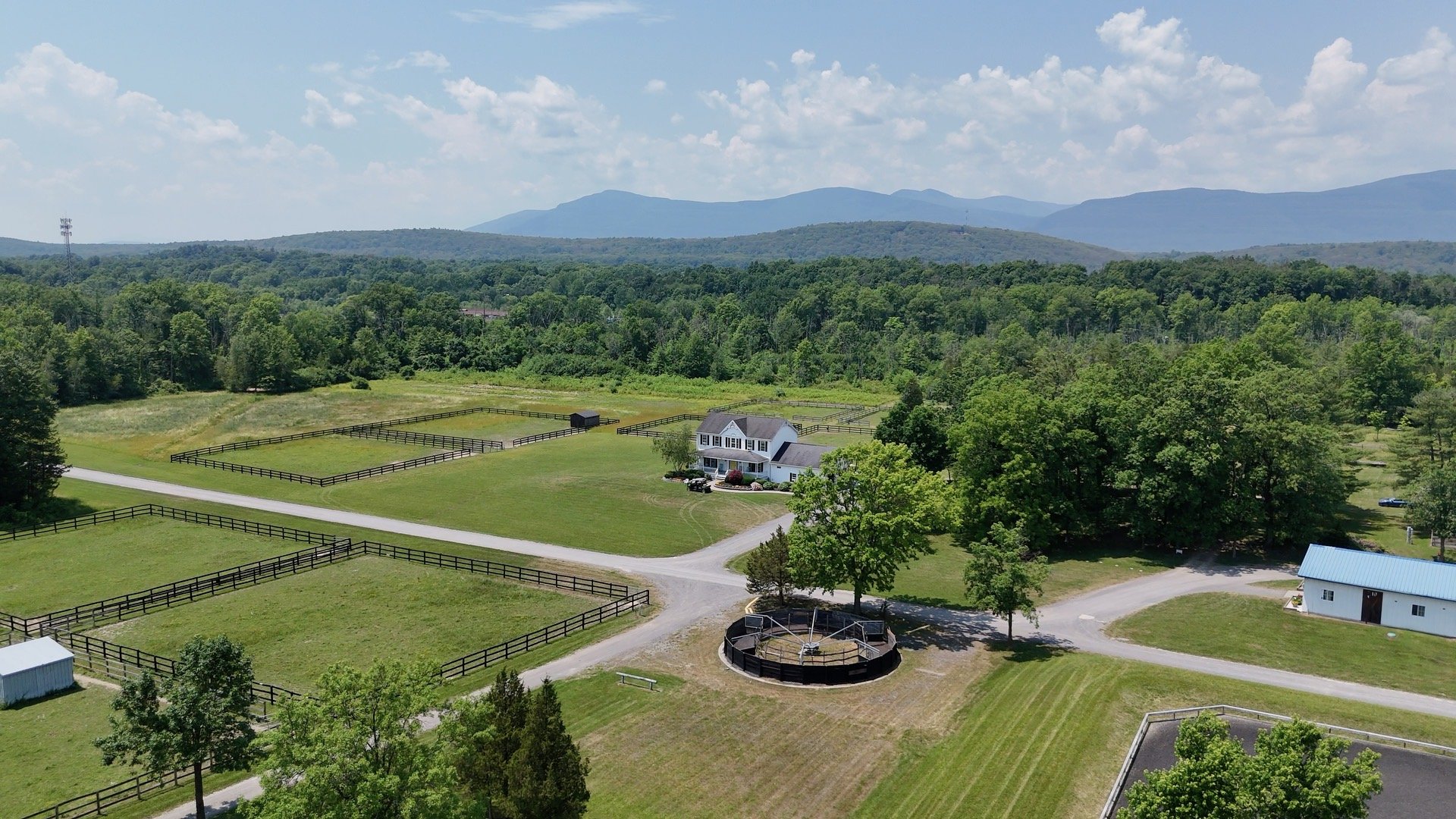 Aerial view of a rural property with a white house, fenced paddocks, a round pen, trees, and distant mountains.