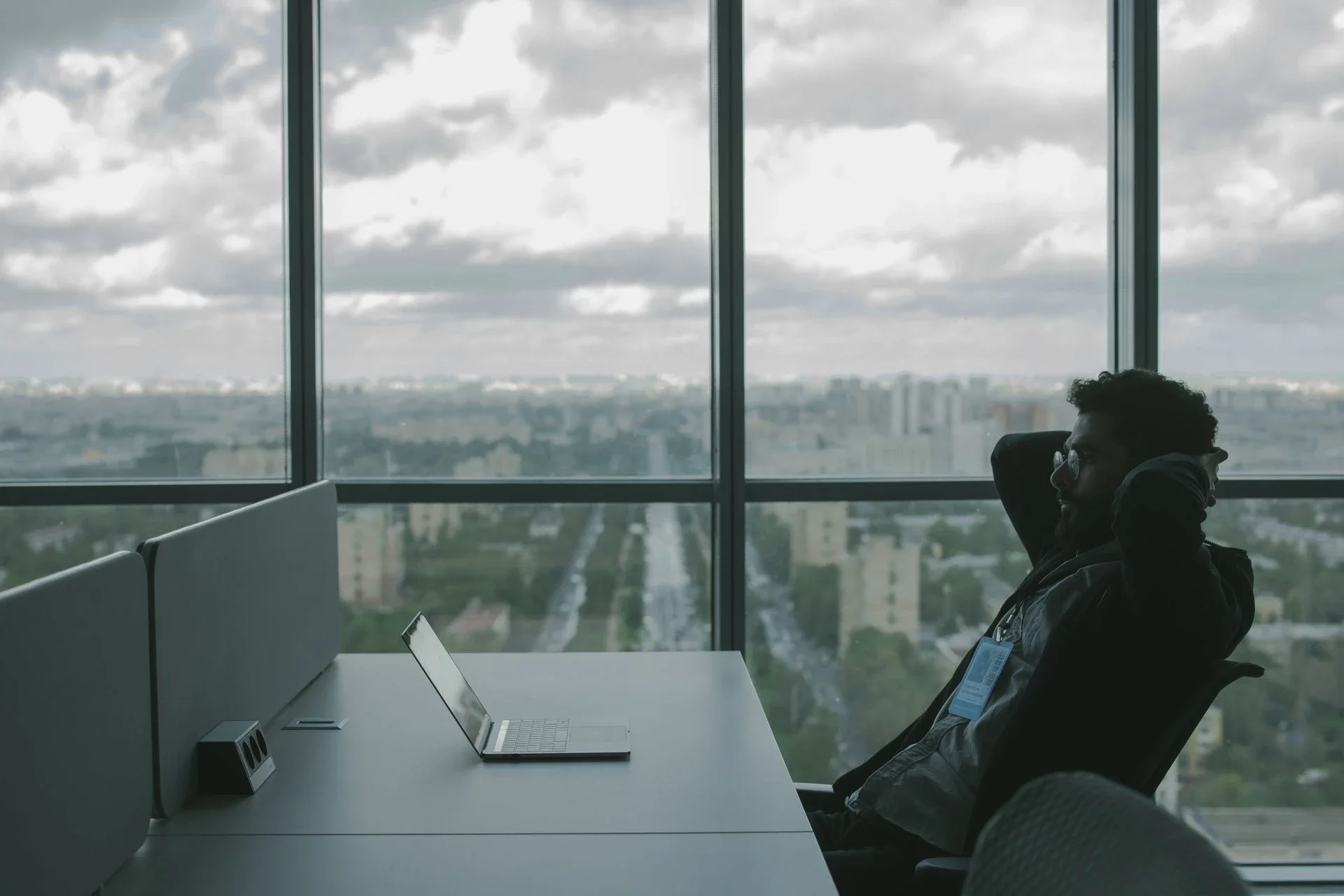 A man sits in an office with hands on his head in front of a laptop, overlooking a cityscape.