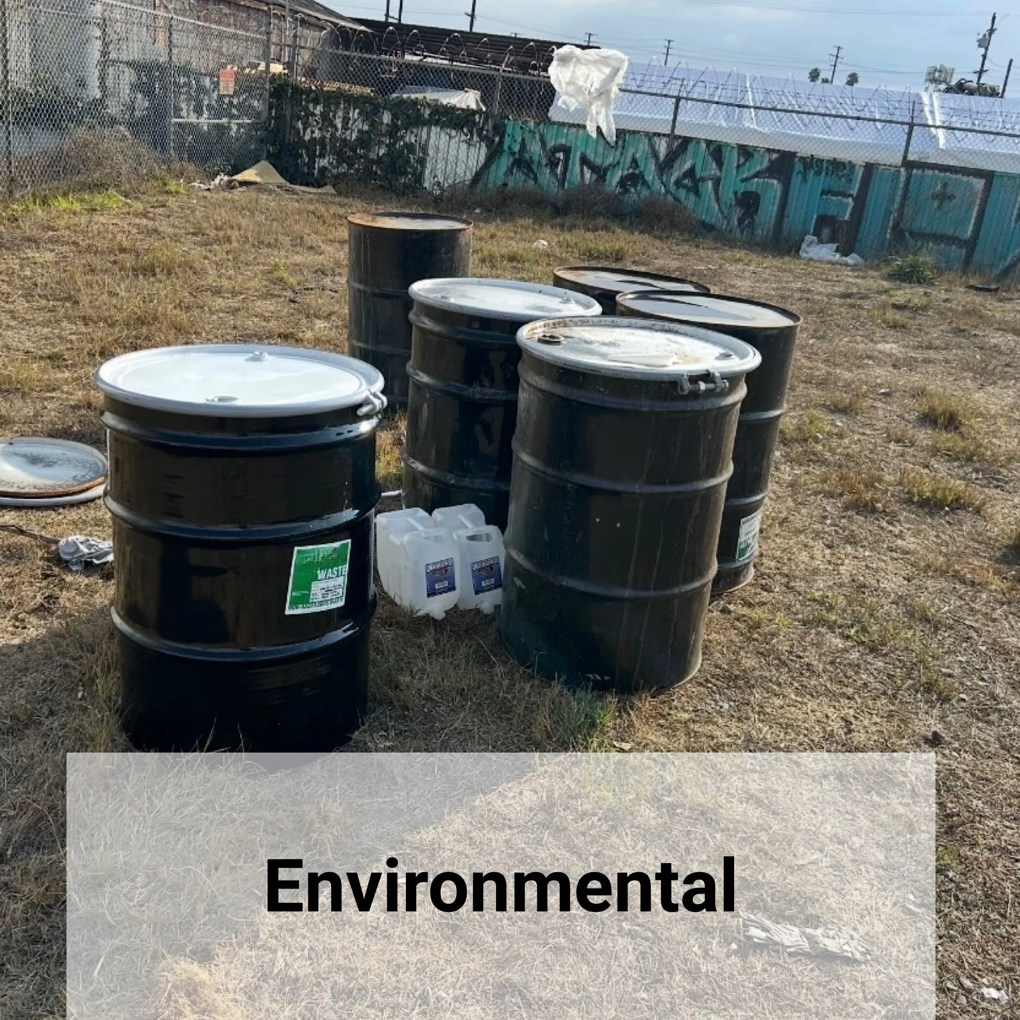 Several black plastic drums with white lids, some stacked and some on the ground, in an outdoor area with dry grass, a chain-link fence, graffiti-covered wall, and trash in the background. The word "Environmental" is overlaid on the image.