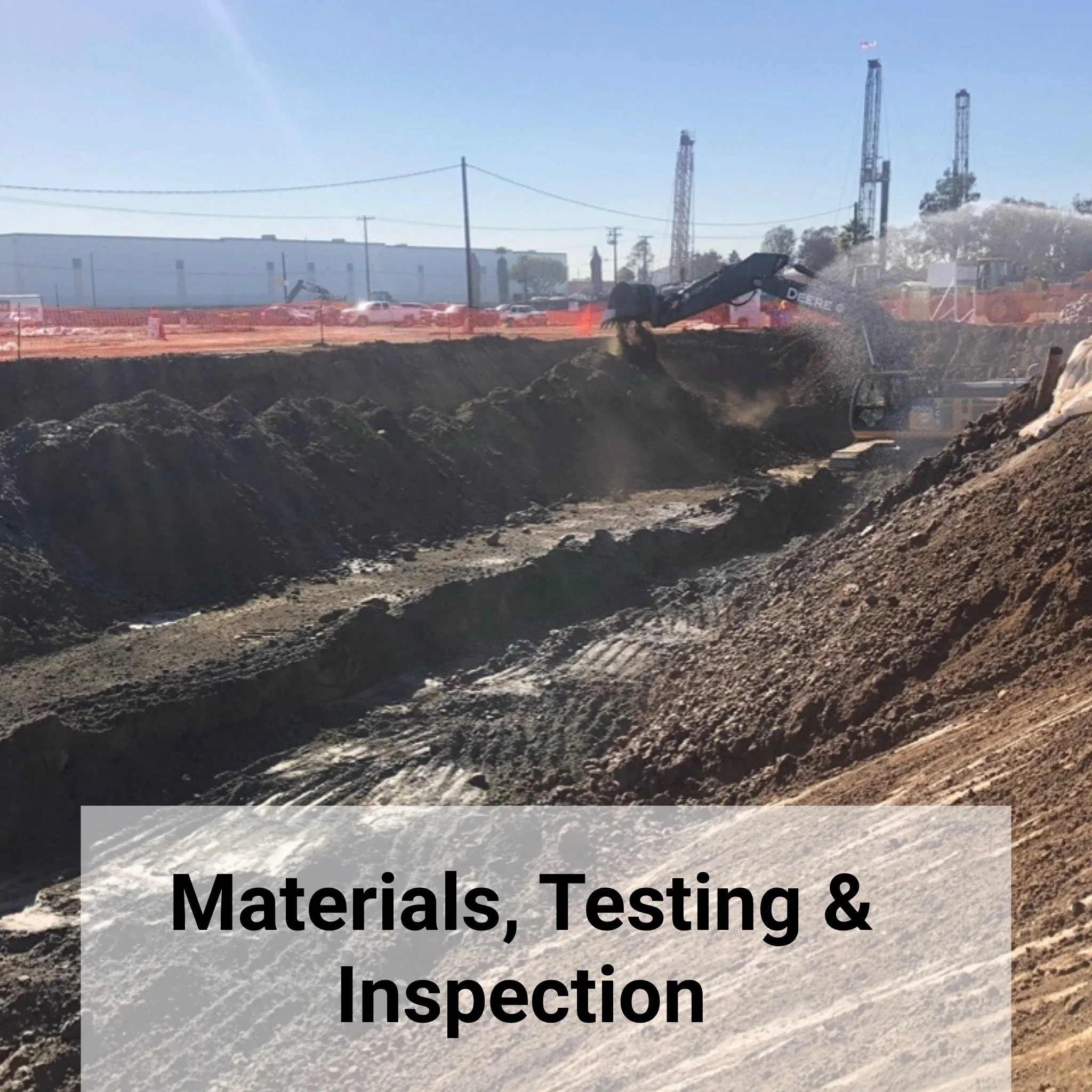 Construction site with excavators digging into the ground, with dirt and soil being moved. Background shows industrial buildings and machinery.