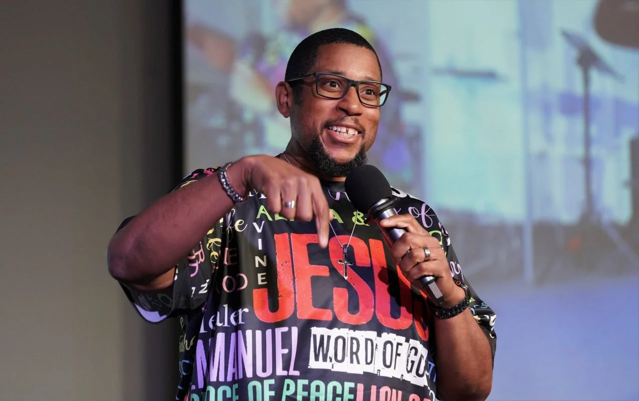 Man speaking during a presentation, wearing glasses, a black T-shirt with text 'JOHN 3:16', and a headset microphone.