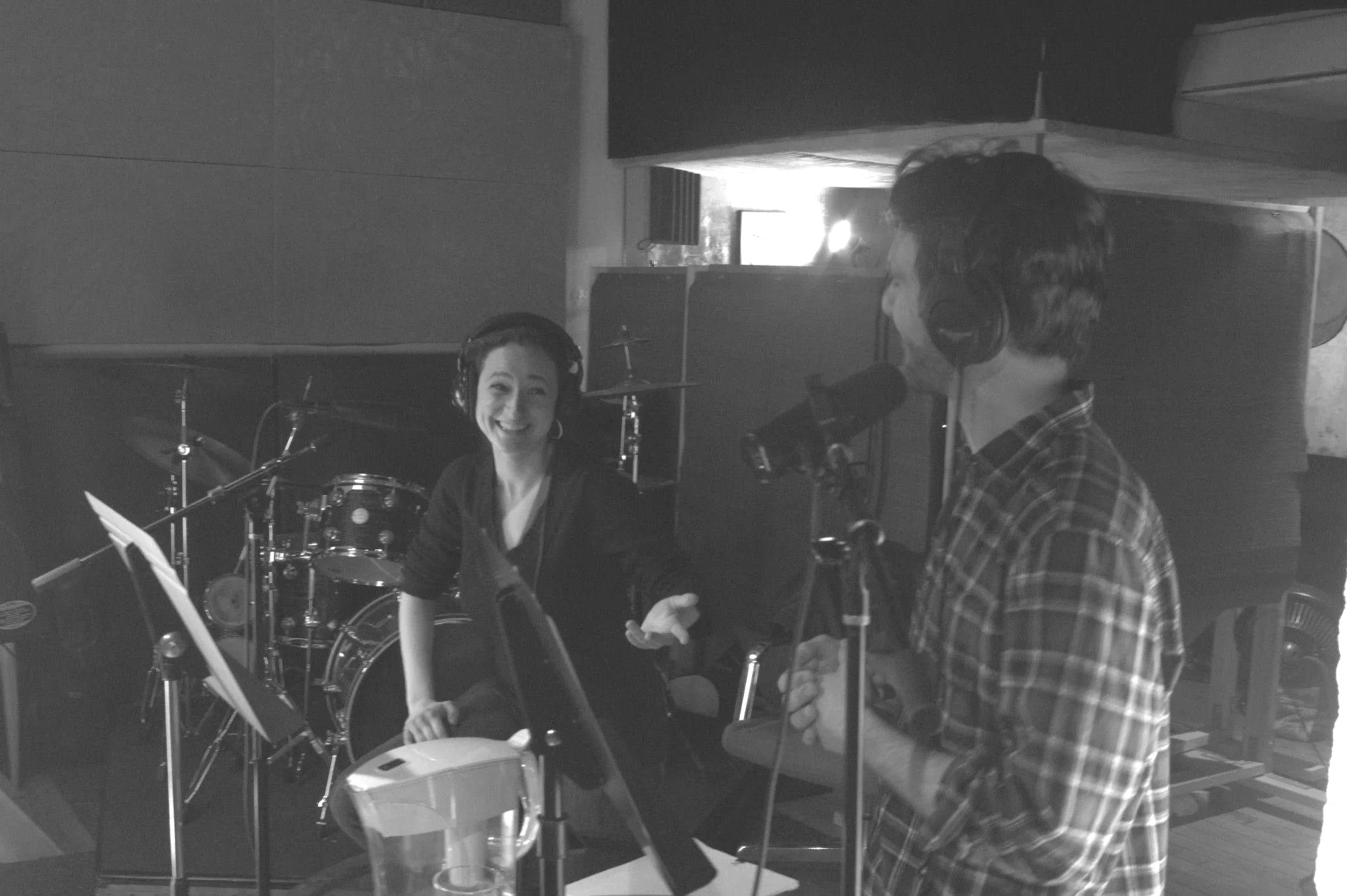 A black and white photo of two people in a recording studio. A woman is sitting and smiling at the other figure who stands and looks back at her.