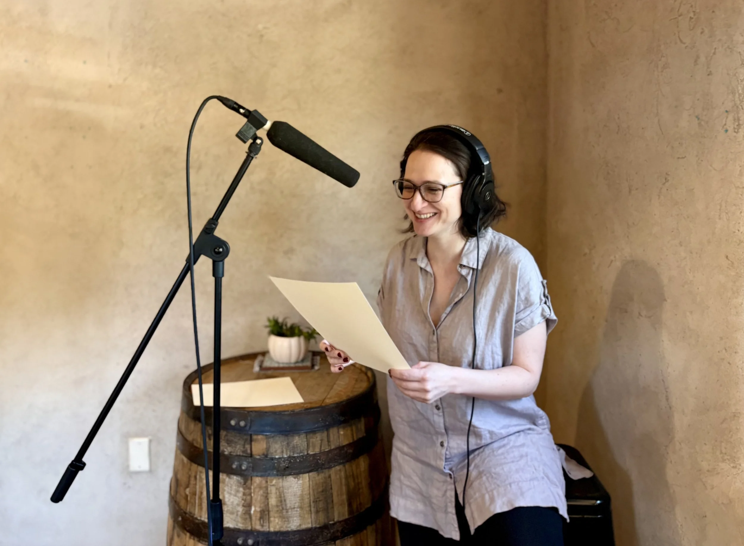 A photo of a woman sitting at a barrel with a microphone. She holds a paper and is smiling.