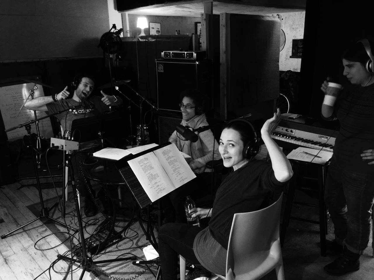A black and white photo of 3 people sitting with music stands and scripts in front of them. They are smiling at the camera. Another figure stands to the right about to drink from a to-go cup.