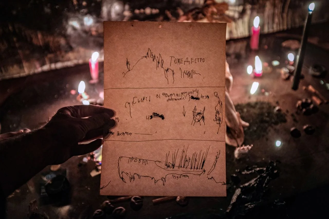 A person's hand holding a handwritten note with drawings and text in front of a lit candle arrangement, burning candles, and ashes on a dark surface.