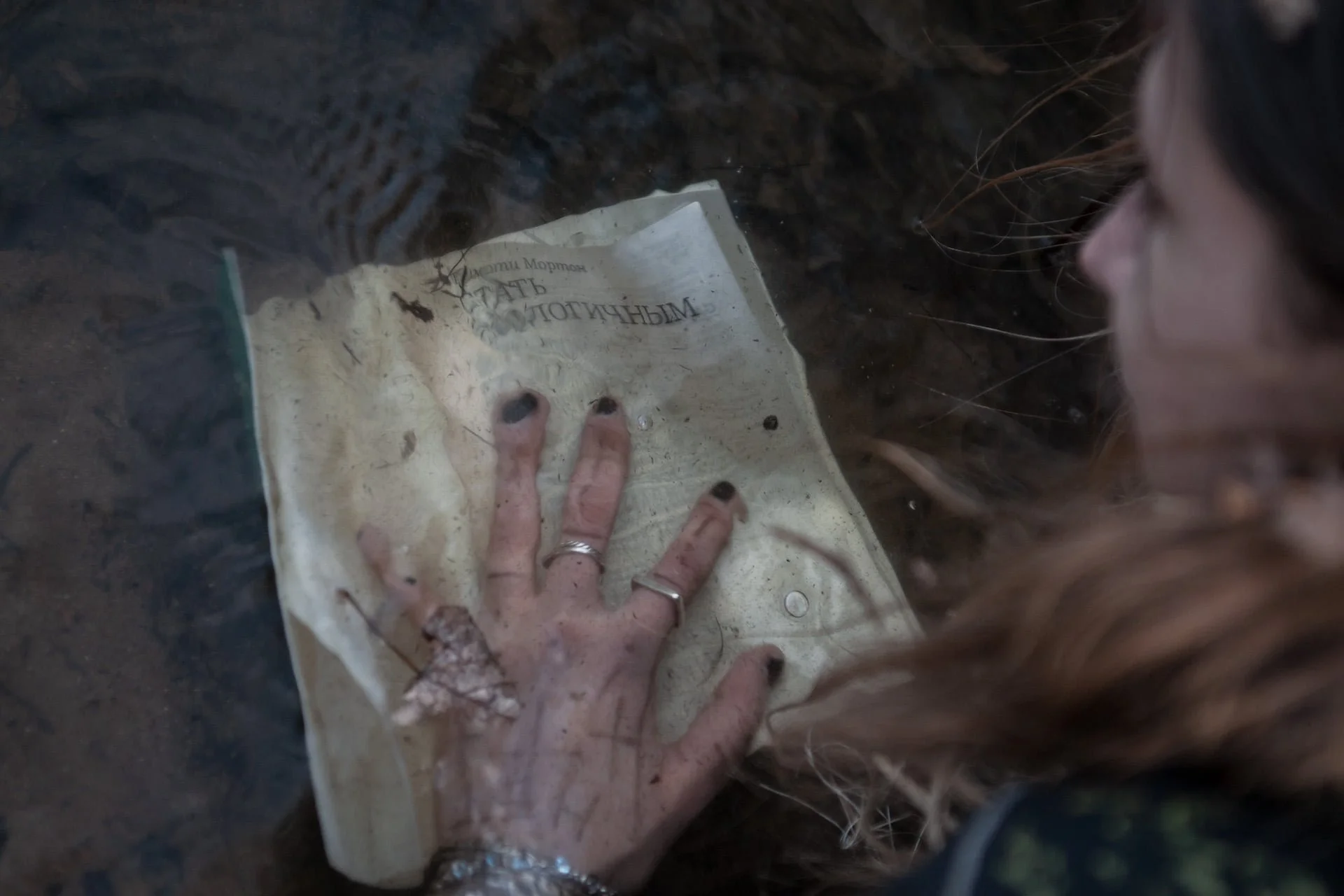 A woman with painted black nails and silver jewelry touching an old, dirty book with Russian text, submerged in water in a muddy environment.