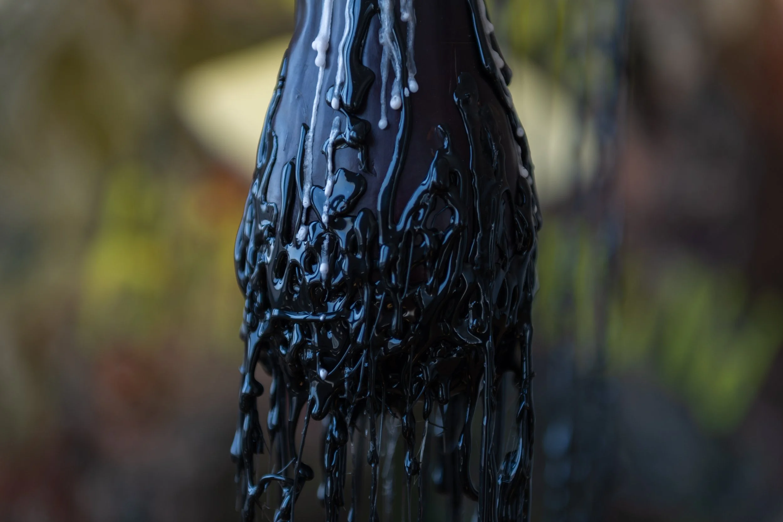 Close-up of a dark, elongated object covered in thick, black, glossy, dripping substance, possibly oil or paint, with a blurred natural background.