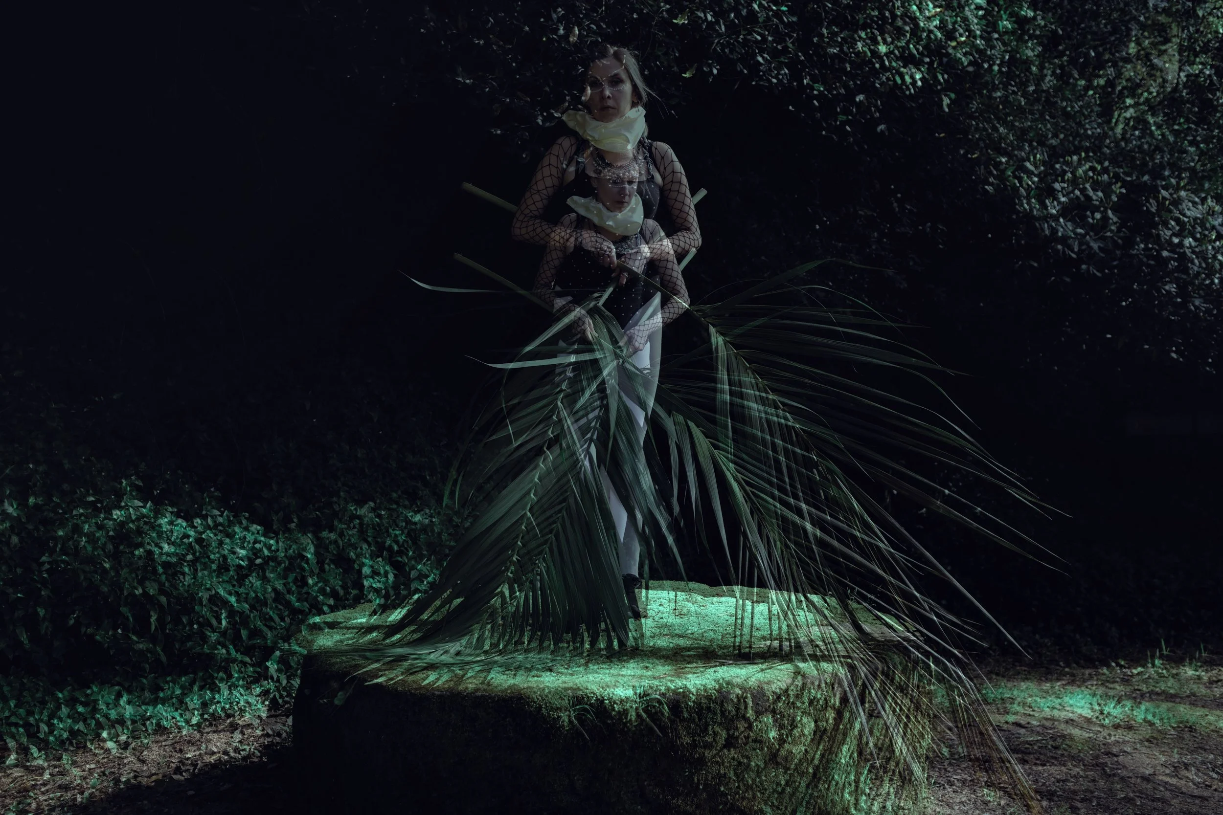 A woman and a girl lying on a bench in a dark, forested area with large leaves and greenery around them.