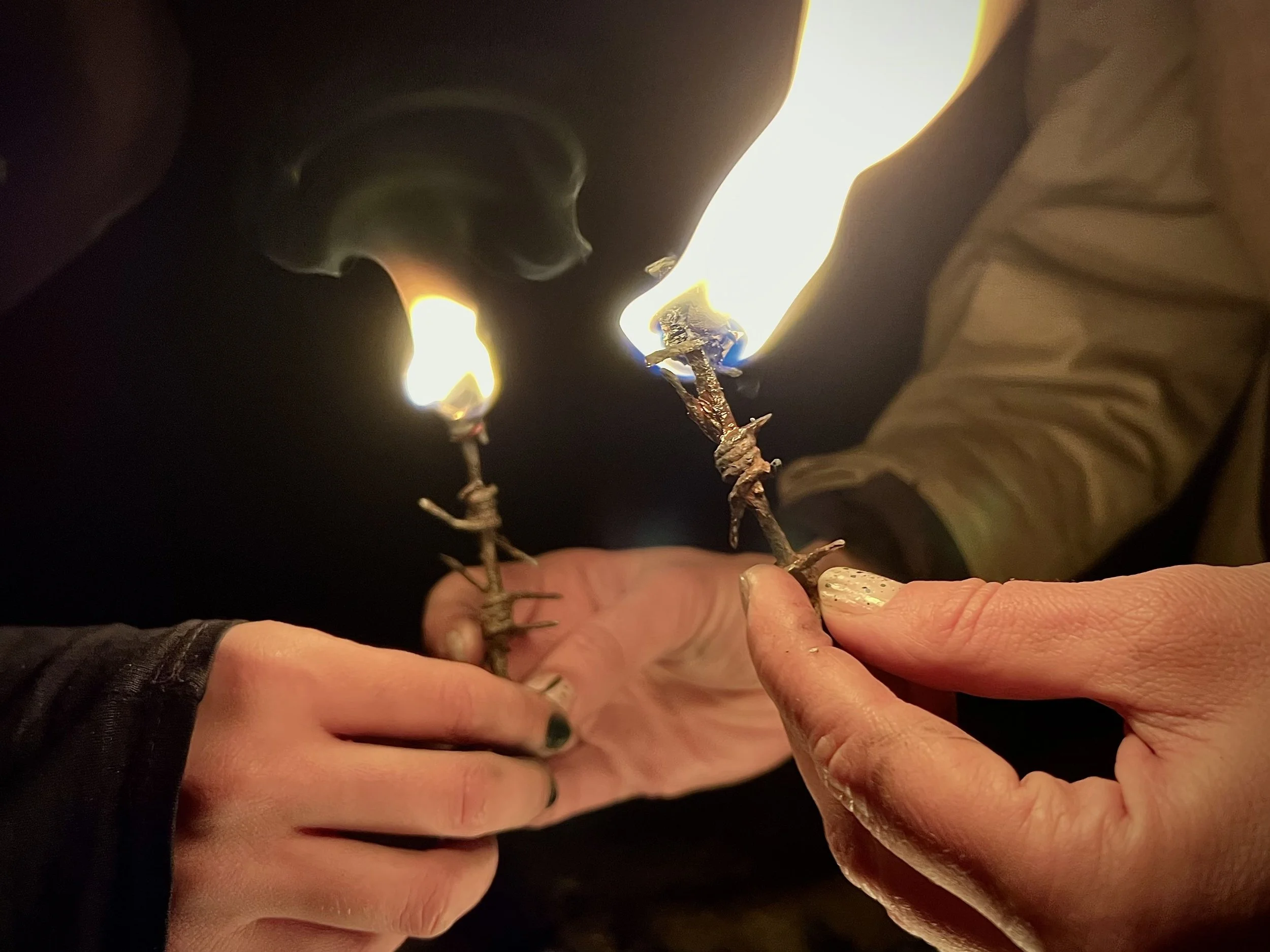 Two people holding small sticks with lit flames at the top, which are wrapped in barbed wire, against a dark background.