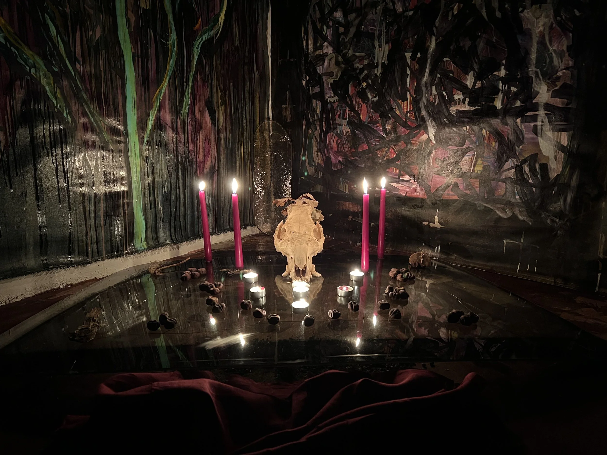 A dark richly decorated altar with a central animal skull, surrounded by four lit candles with pink holders, small candles, scattered beans or seeds, and a dark, abstract, colorful background on the wall.