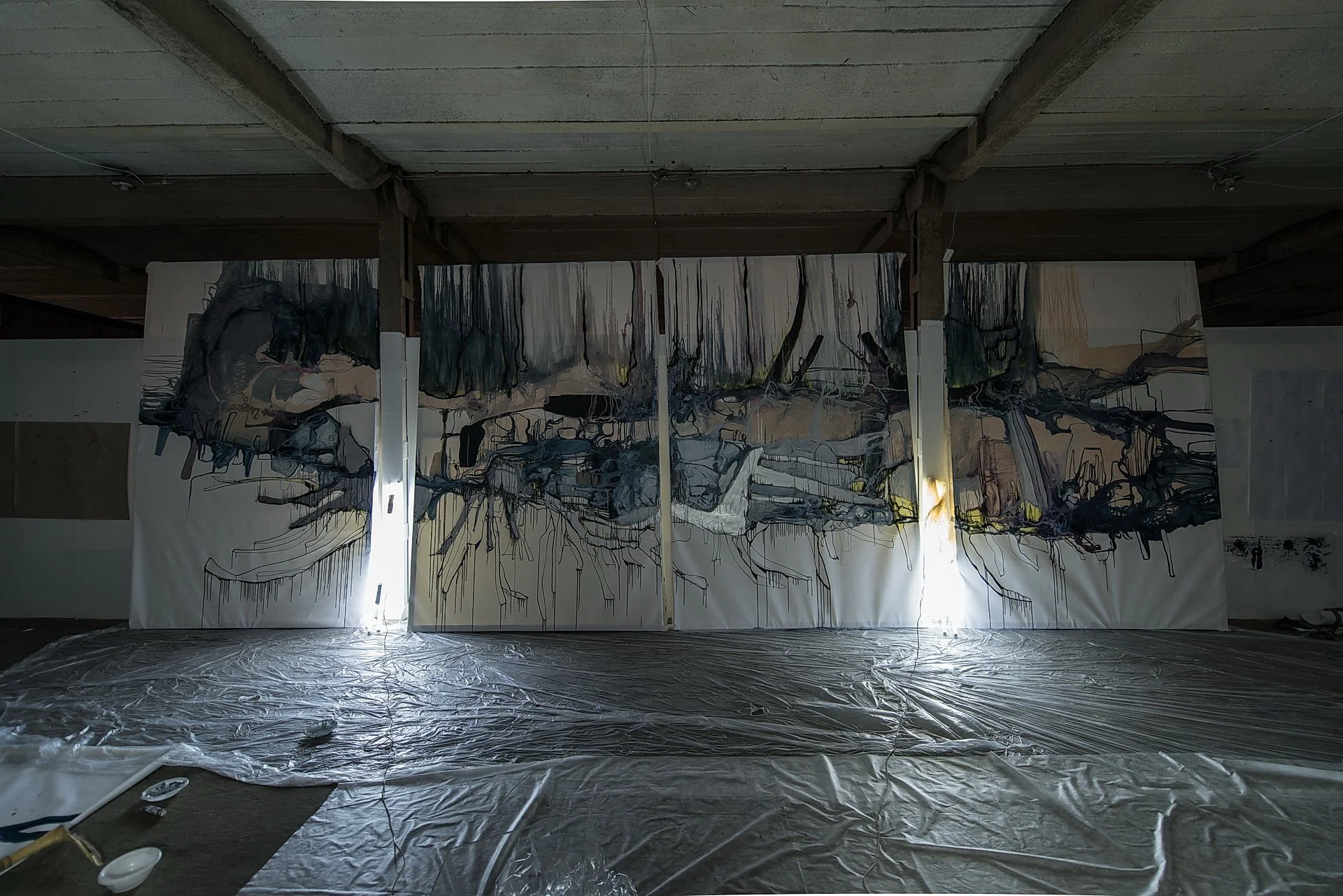 Large abstract painting with dark, fluid lines and muted colors on a multi-panel canvas, held in a spacious room with plastic covering on the floor and ceiling.