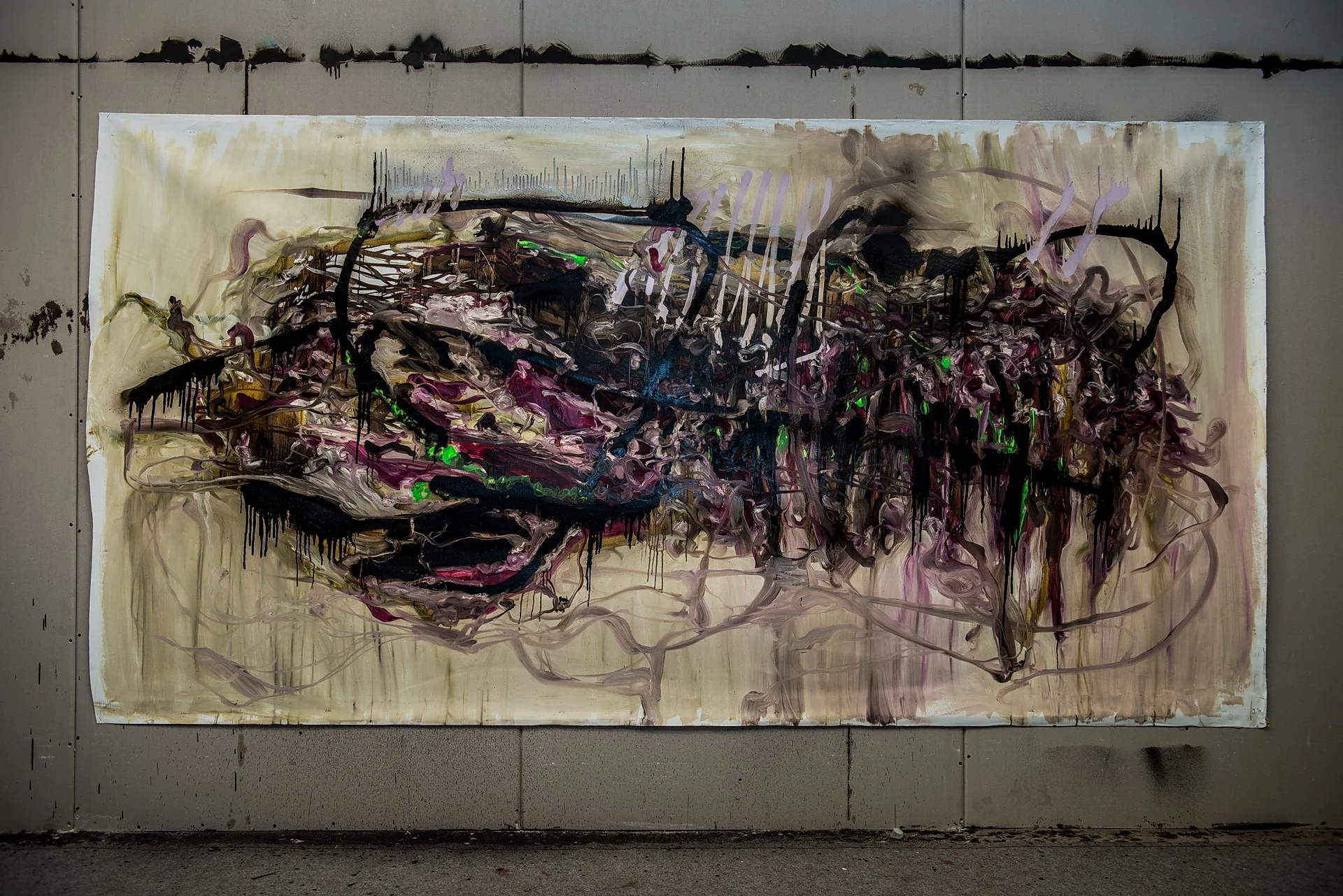 An abstract painting on a large canvas mounted on a gray wall, featuring chaotic swirls of black, purple, white, and green colors with drips and lines creating a complex, expressive composition.