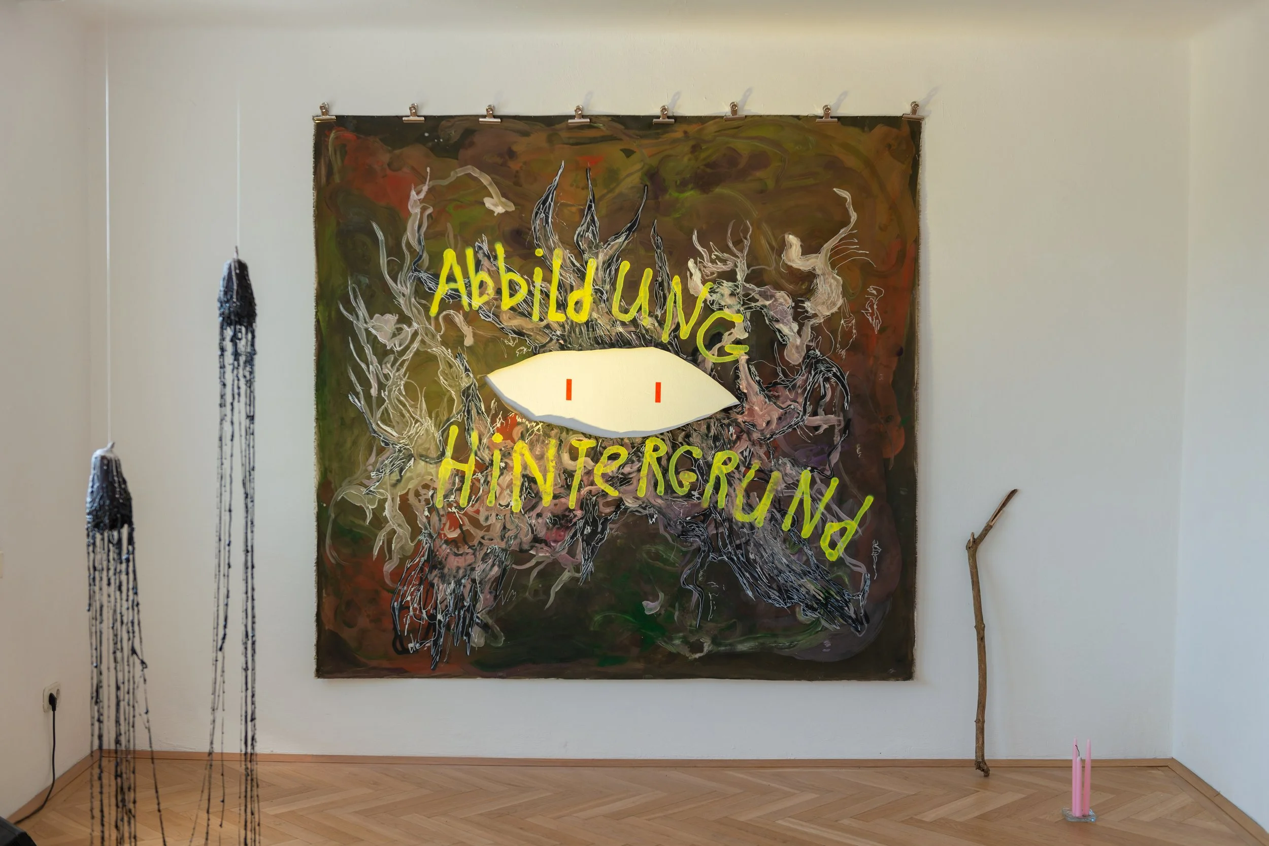 Mixed media art installation with a colorful canvas featuring the phrase 'Abbild und Ahnengruß' and a stylized eye in the center, surrounded by abstract shapes and lines, with hanging black and blue beads on the left, a wooden stick on the right, and