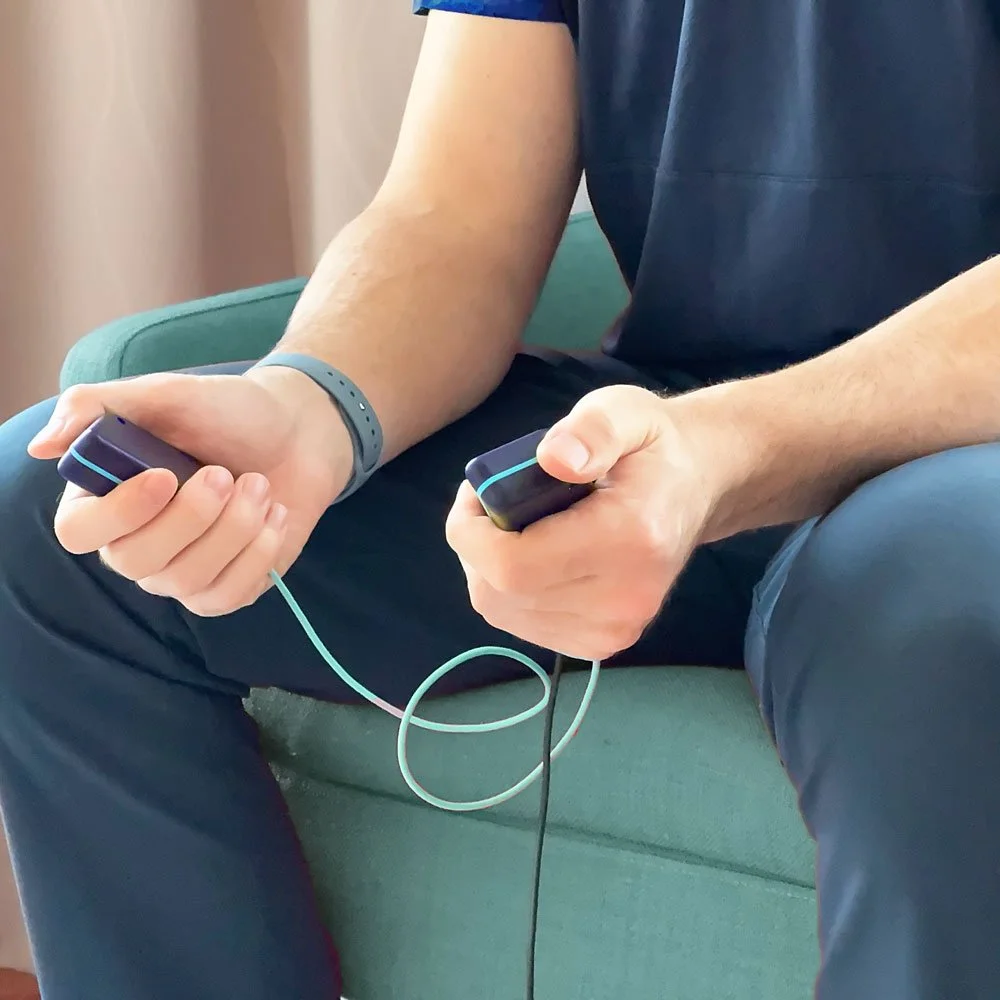 Person sitting on a green armchair, holding a device in each hand, with wires connected to their wrists. The person is wearing a dark shirt and dark pants, and a gray wristband on one wrist.