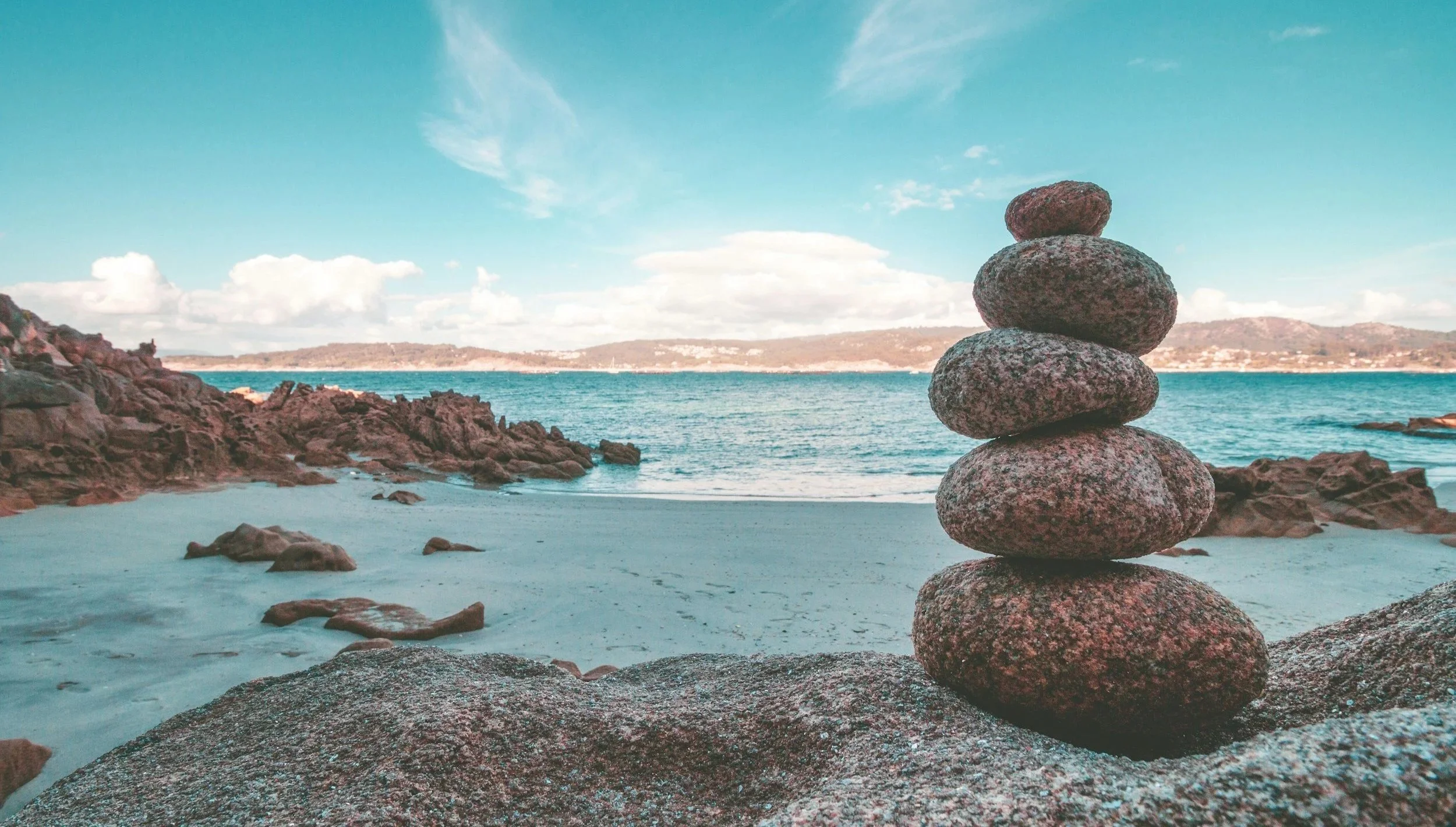 A stack of seven rounded rocks balanced on a beach with ocean water, rocky formations, and a distant landmass under a partly cloudy sky.