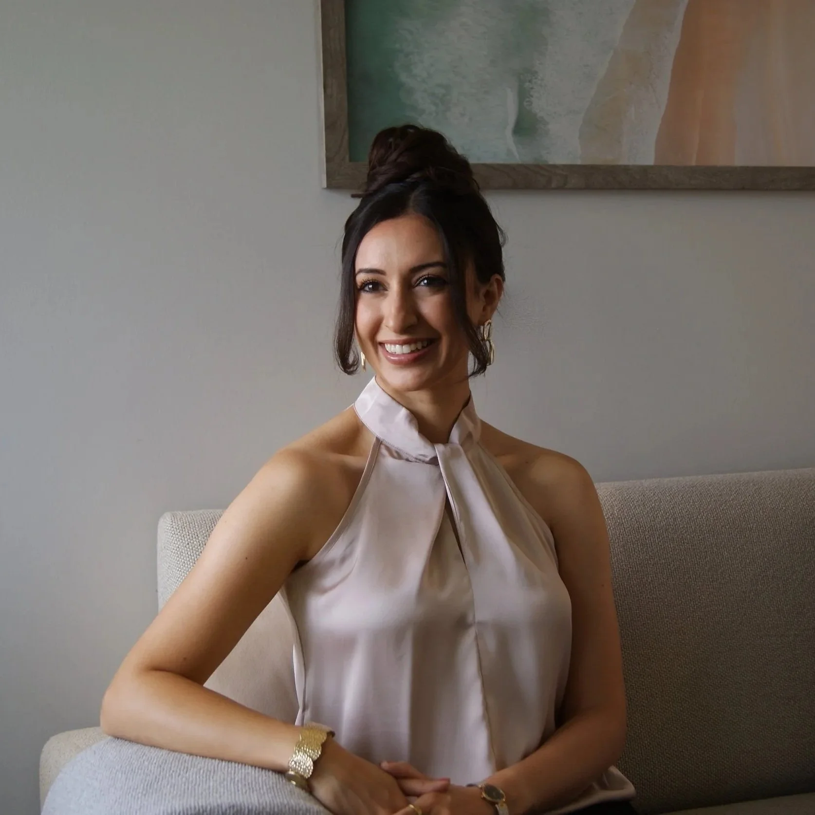 A woman with dark hair styled in a top bun, smiling and sitting on a beige couch in front of a plain light-colored wall with a framed abstract artwork hanging above her. She is wearing a light satin halter top and gold jewelry, including earrings, a bracelet, and a watch.