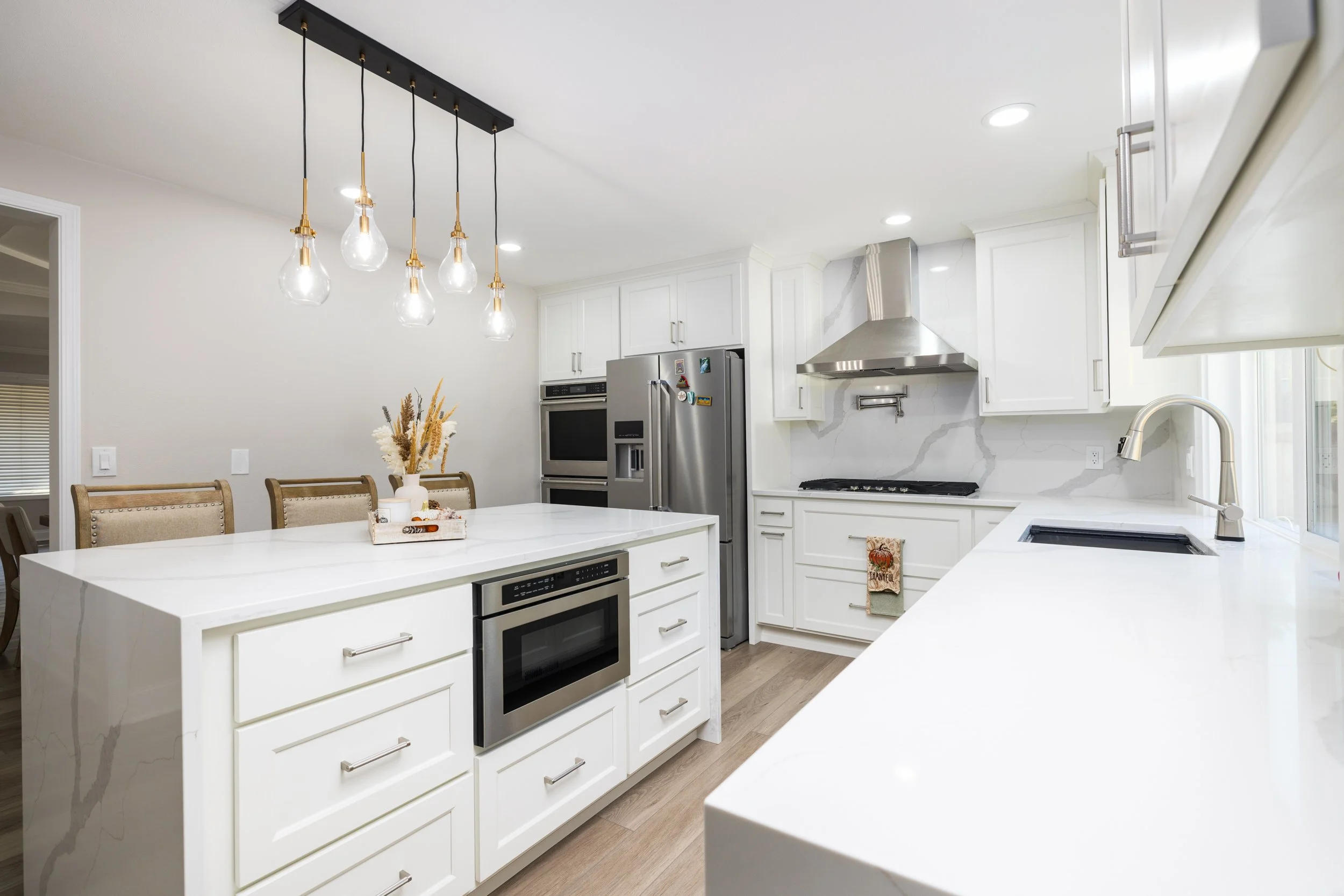Modern kitchen with white cabinets, marble backsplash, stainless steel appliances, and a white central island. Pendant lights with exposed bulbs hang from a black fixture. There are wood chairs with beige upholstery around the island. Natural light e