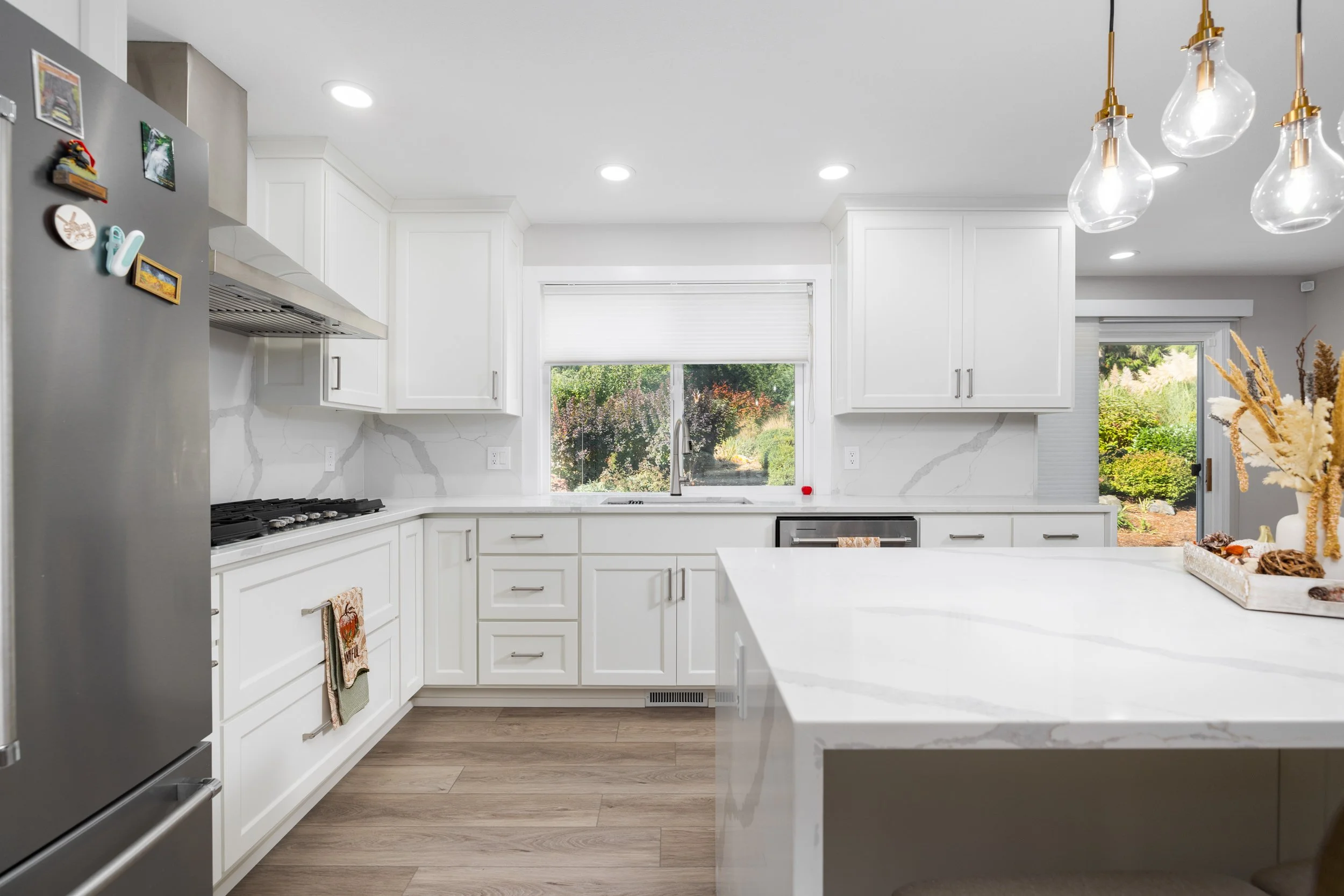 A modern kitchen with white cabinets, marble countertops, a large window above the sink, pendant lights, and a stainless steel refrigerator with magnets, against a window with a view of a garden.