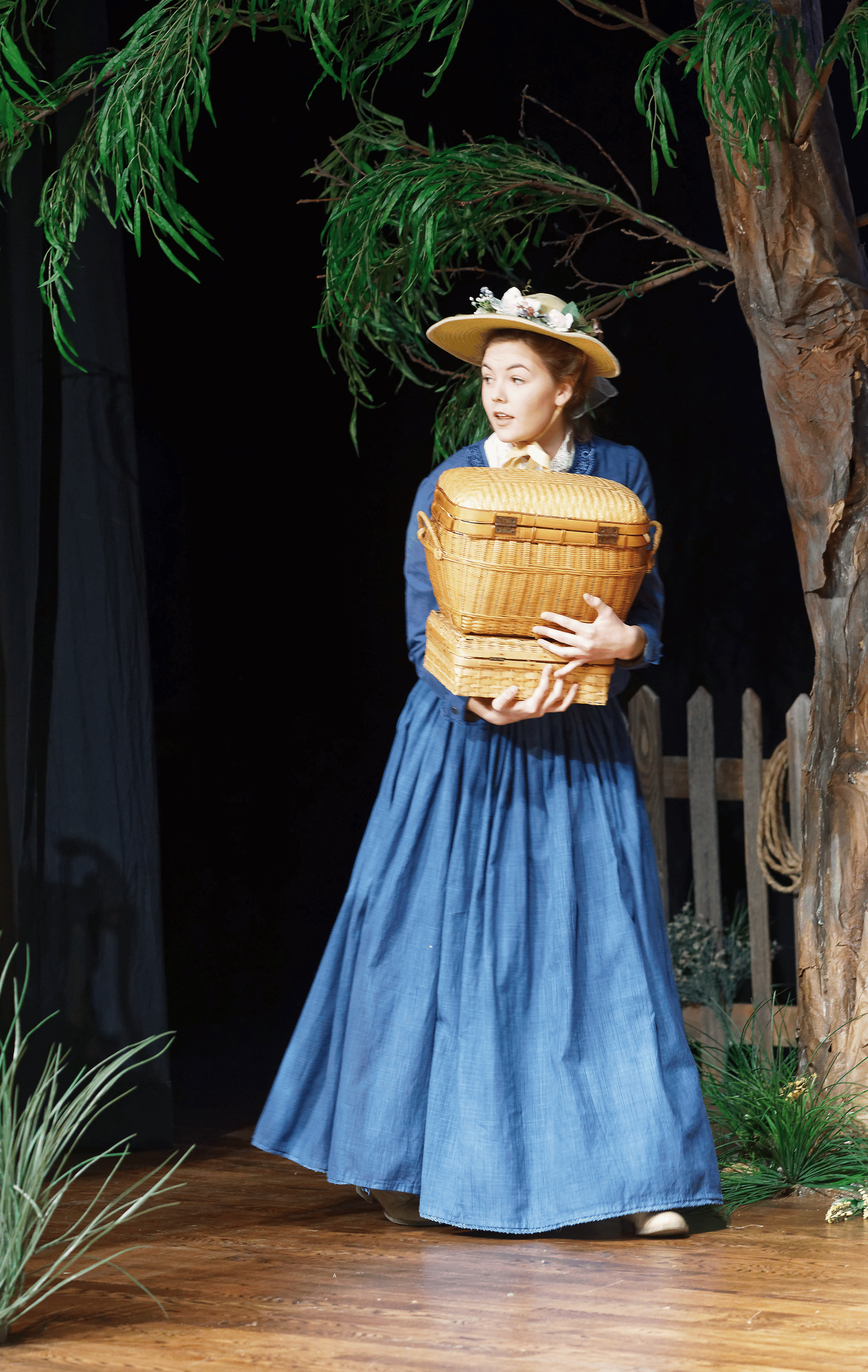 A woman dressed in historical costume, wearing a wide-brimmed straw hat decorated with flowers, and a blue long dress, holding a wicker basket, standing on a wooden stage with a naturalistic set resembling a garden or outdoor scene.