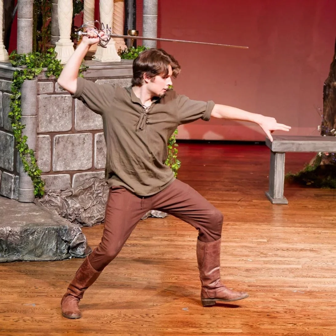 A young man dressed in medieval-style brown tunic, pants, and tall boots practicing fencing indoors with a sword, in front of a faux castle backdrop.