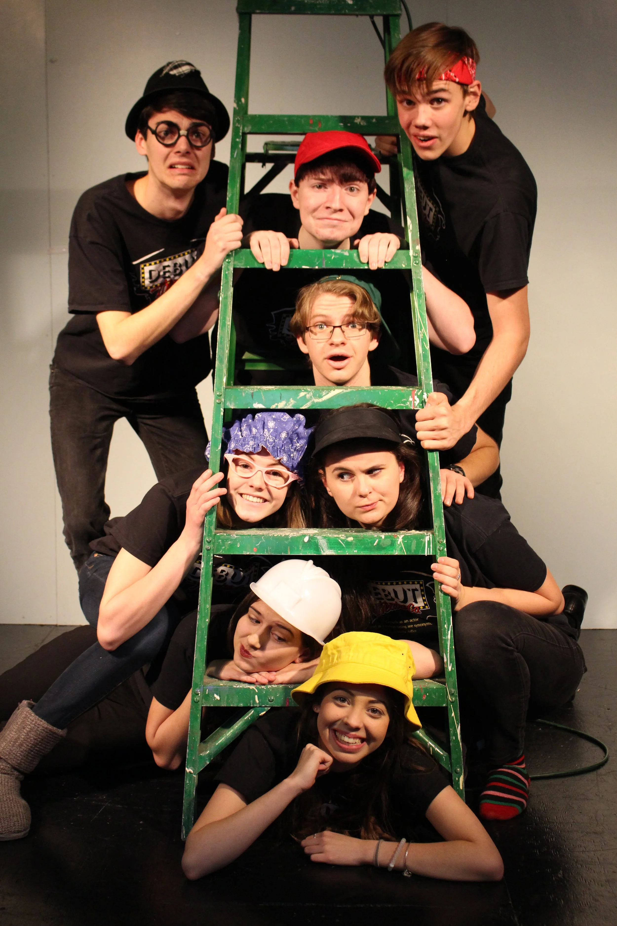 Group of people stacking on a green ladder, dressed in costumes and accessories, posing on a stage with a plain background.