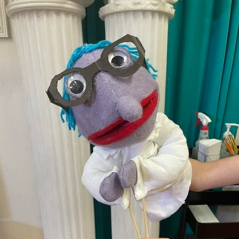 A puppet with purple skin, big googly eyes, blue yarn hair, glasses, red lips, wearing a white coat, held by a person's arm in a room with white columns and teal curtains.