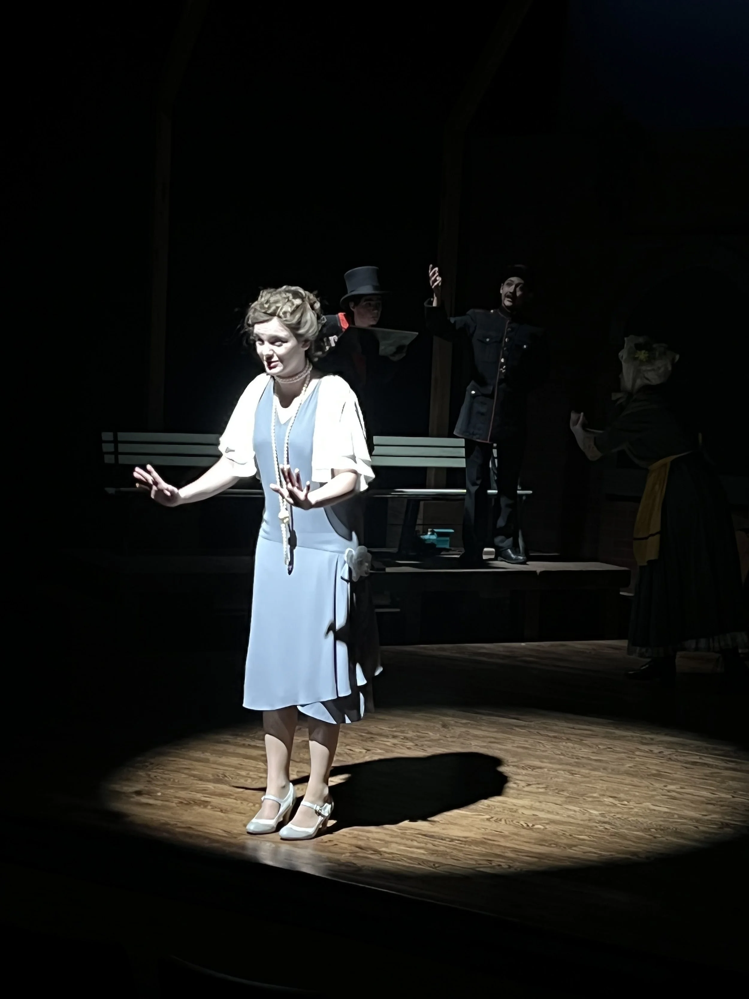 A theatrical performance with actors on stage, including a woman in a light-colored dress and heels, and other actors in dark costumes, illuminated by a spotlight in a darkened theater.