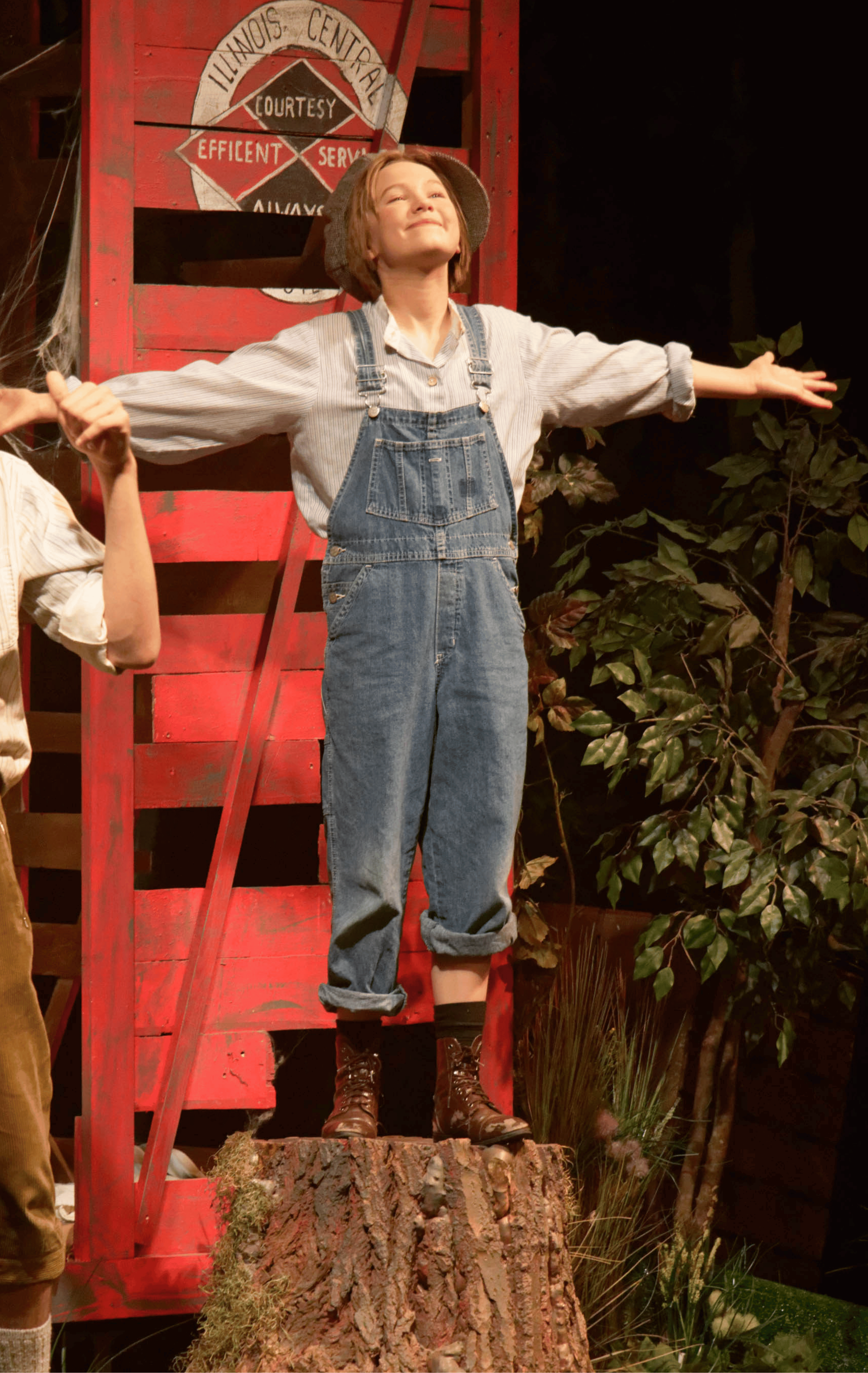 A young woman with short red hair and a cap, wearing overalls and boots, stands on a tree stump with arms outstretched, smiling contentedly. She is in front of a red wooden structure with a sign that reads "Illinois Central" and some greenery.