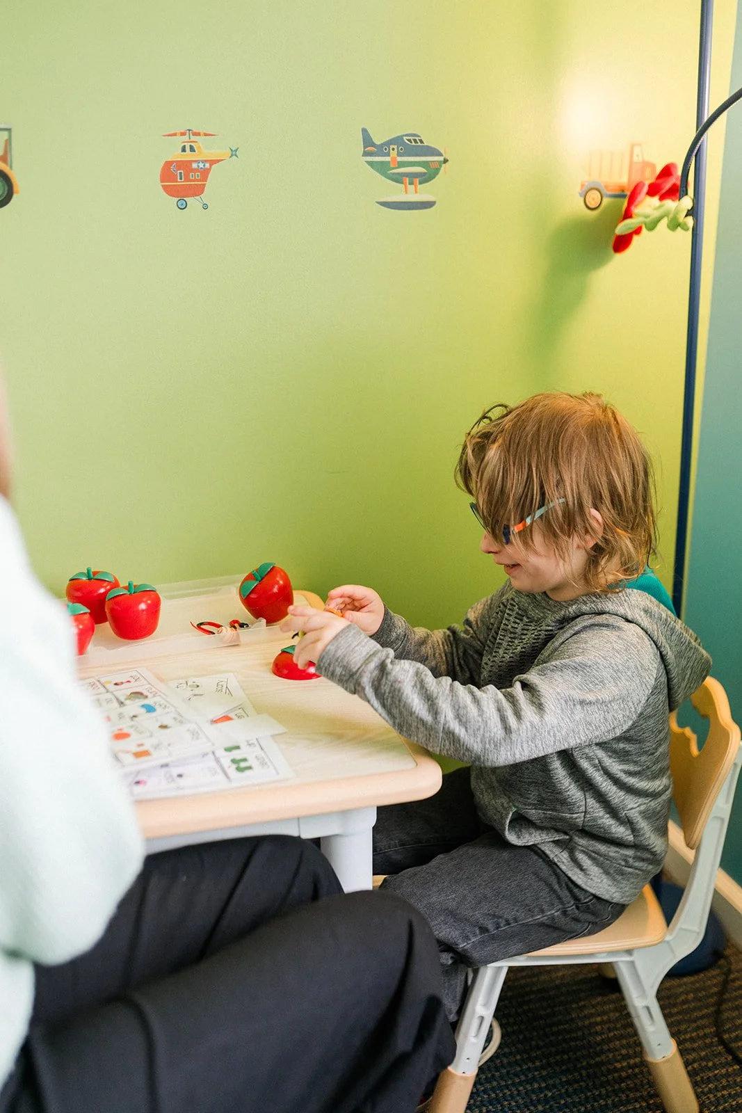 A young boy with glasses, smiling, wearing a gray hoodie, sitting at a small table playing with red apple toys. The background depicts a light green wall with airplane and helicopter stickers, and there are some paper instructions or cards on the table.