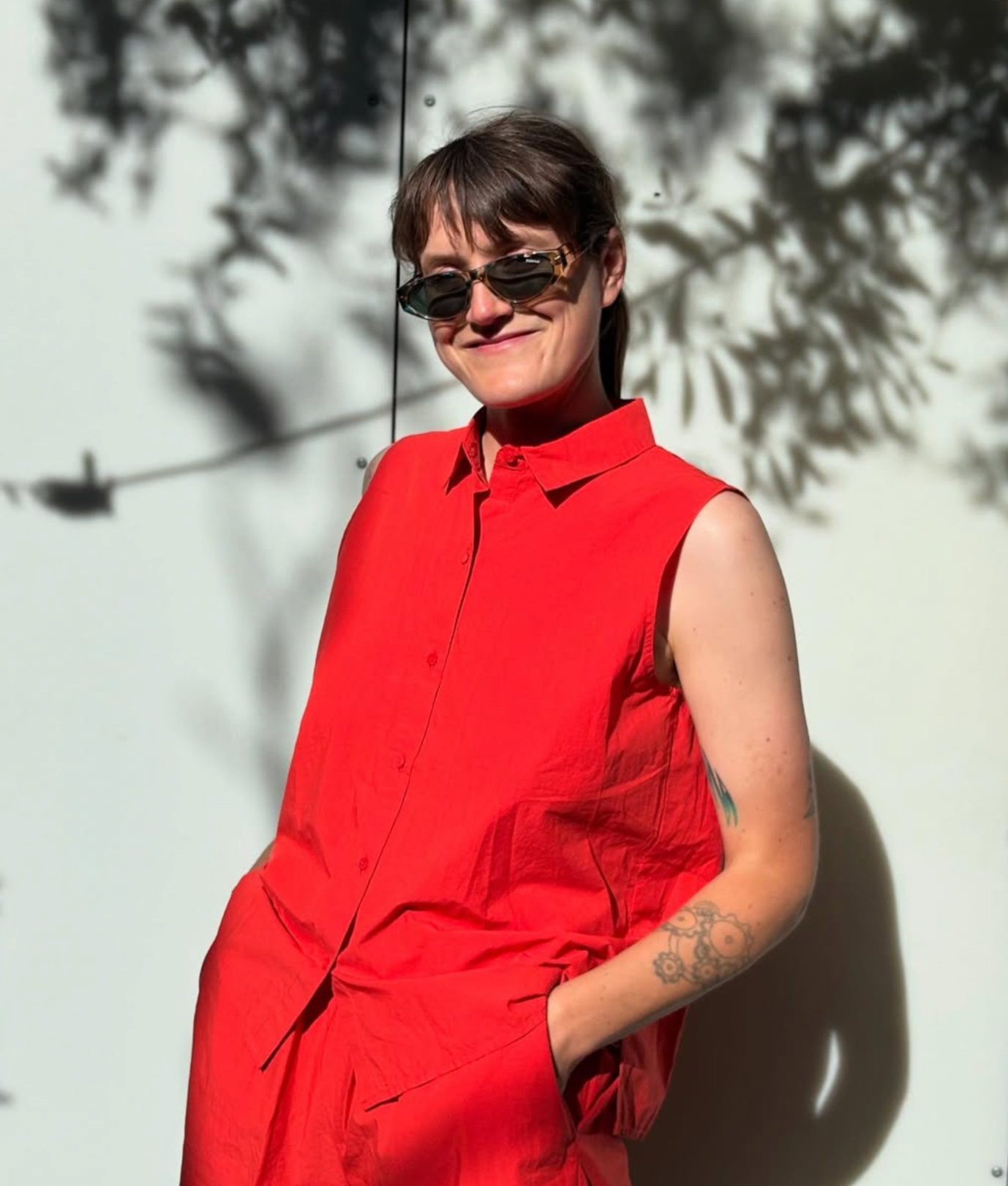 A person with dark hair wearing sunglasses and a sleeveless red shirt standing against a white wall with shadows of tree branches and leaves