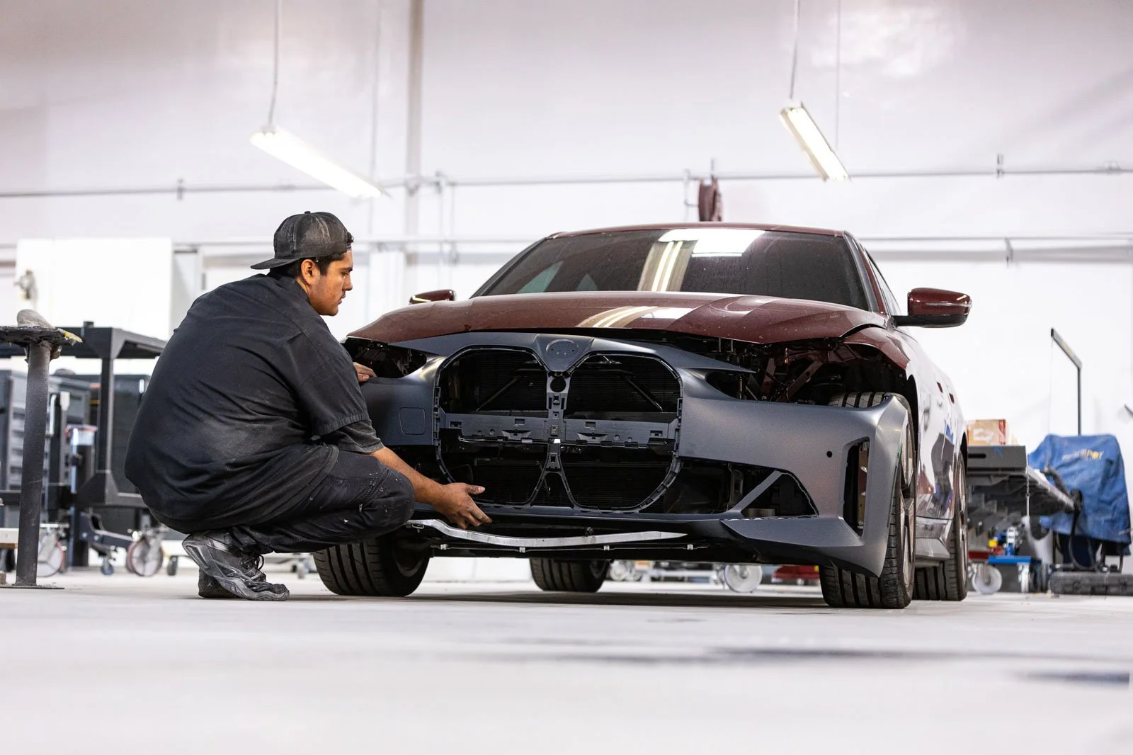 A mechanic kneeling in front of a car undergoing repair in a workshop, working on the front grille area.